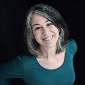 A person smiles warmly, tilting their head slightly, wearing a teal top against a dark background.
