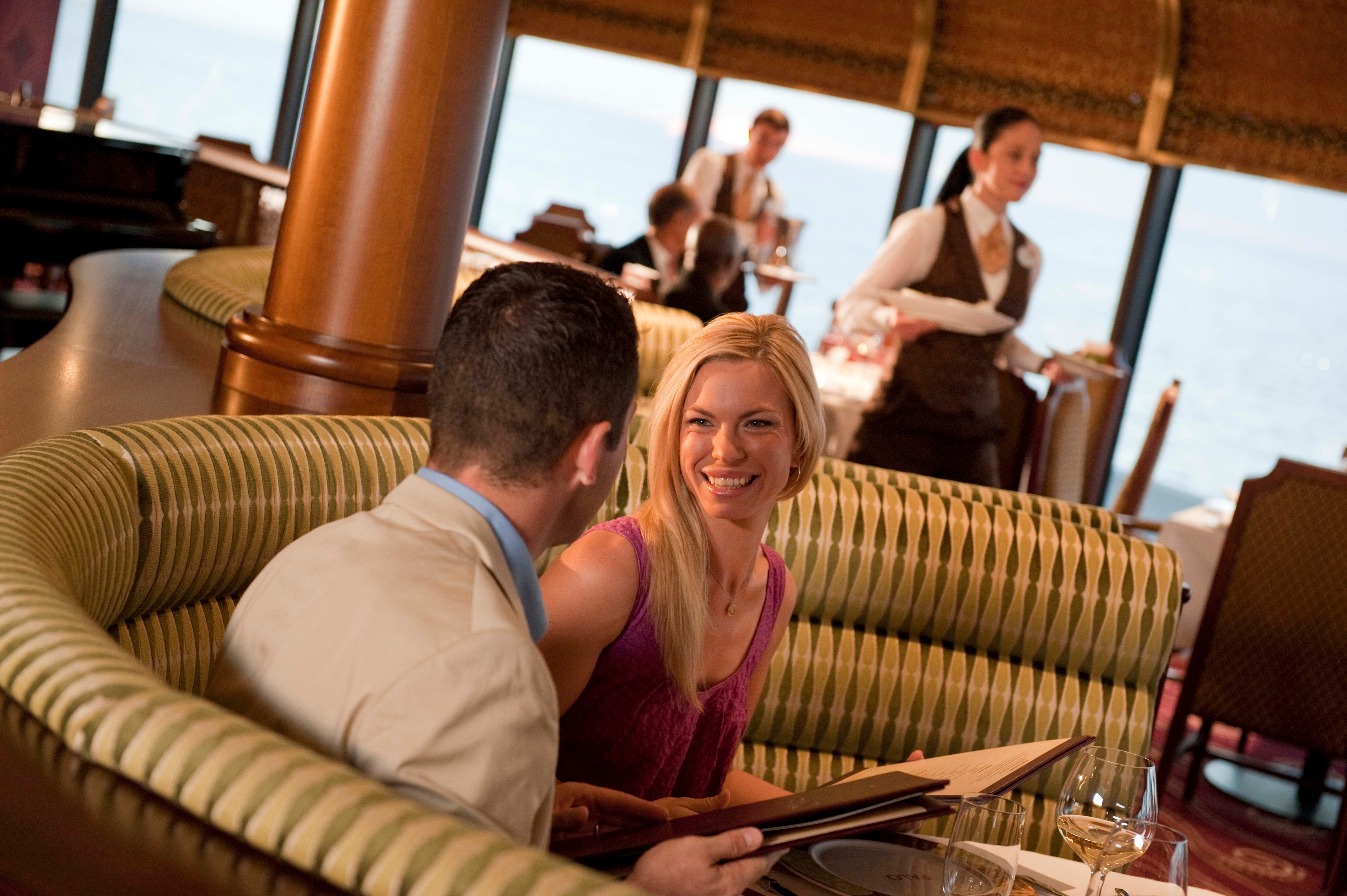 A couple converses at a restaurant, seated in a booth with menus, while waitstaff attend to other guests in the background.
