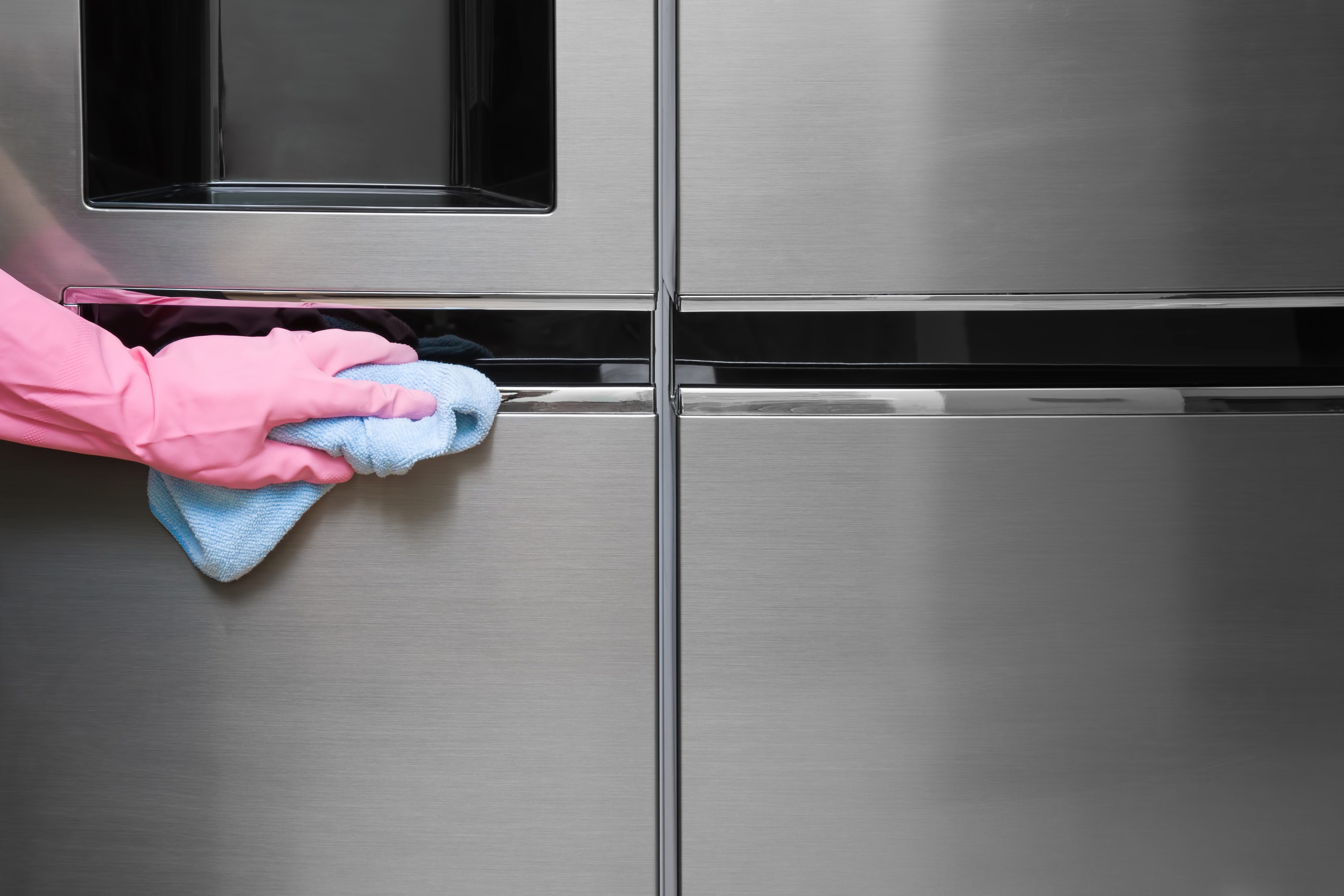 A hand in pink gloves wipes a stainless steel refrigerator door with a blue cloth, highlighting household cleaning.
