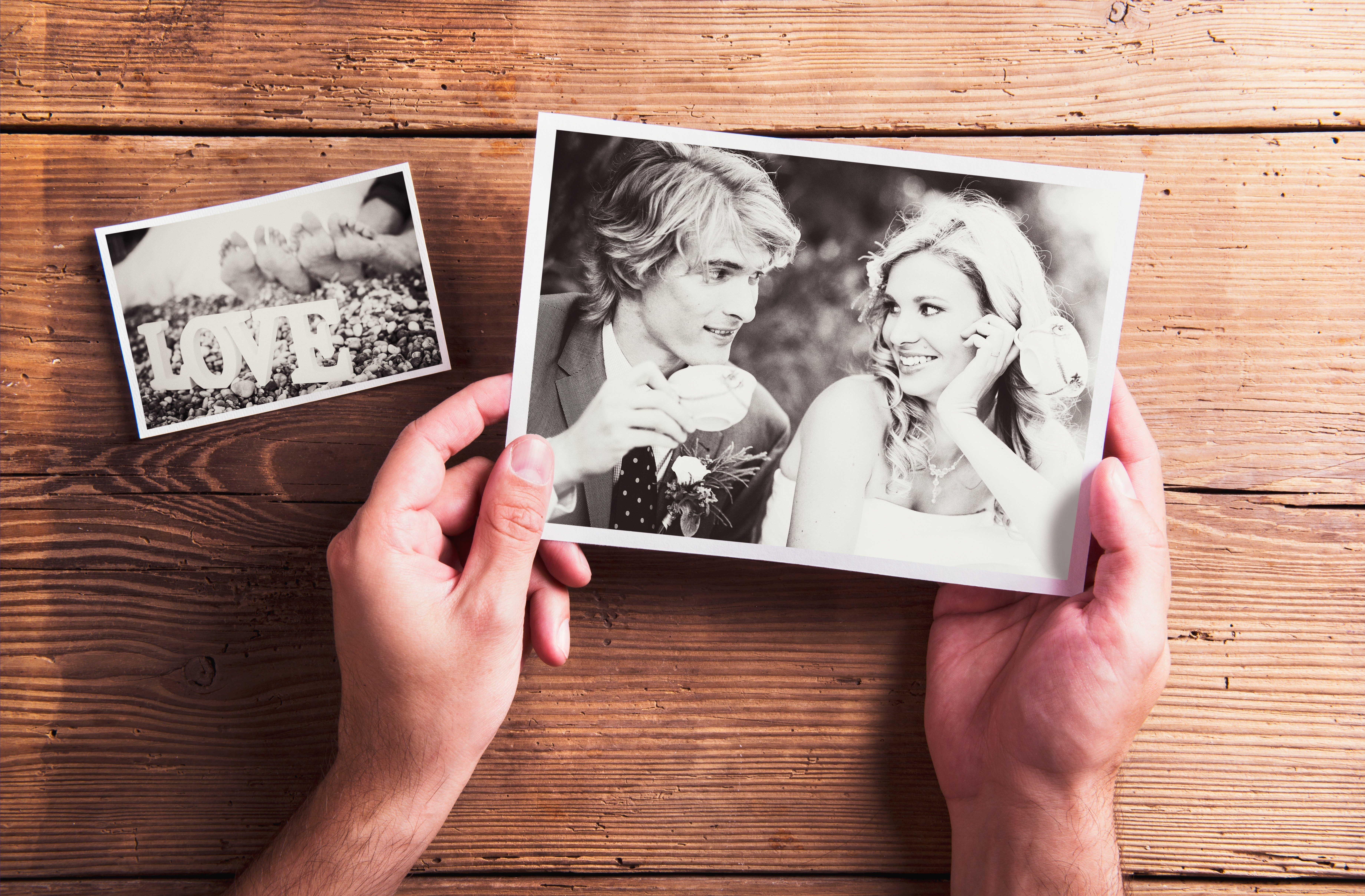 Hands hold a black-and-white photo of smiling couple; nearby, a smaller photo displays bare feet and the word "LOVE" on a wooden table.