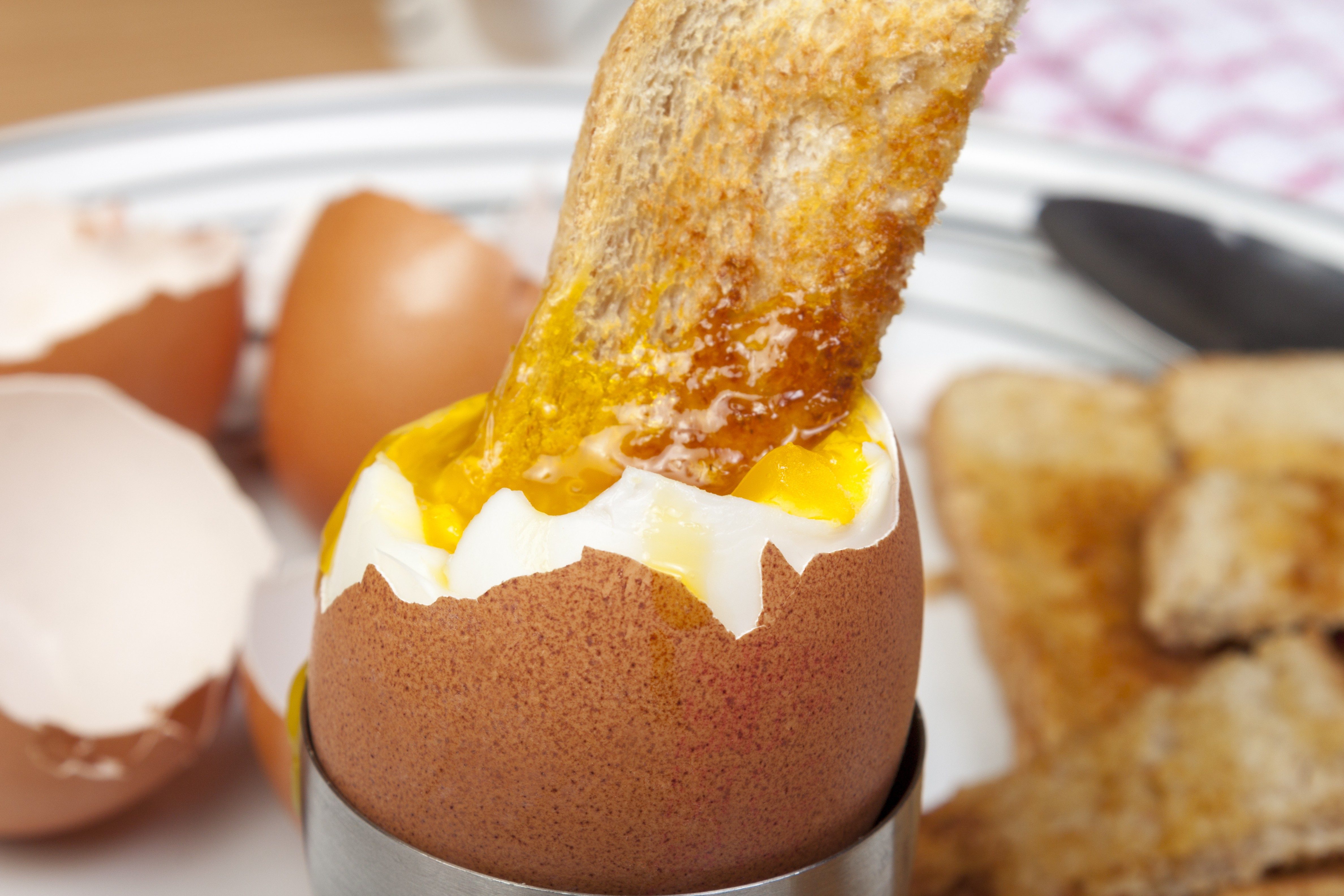 A toast soldier dipping into a soft-boiled egg in an egg cup, with broken eggshells and additional toast slices on the plate.