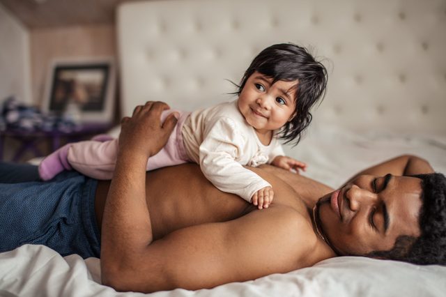 Baby lying on a man's chest, both smiling contently on a bed, with a soft-focus framed picture in the background.