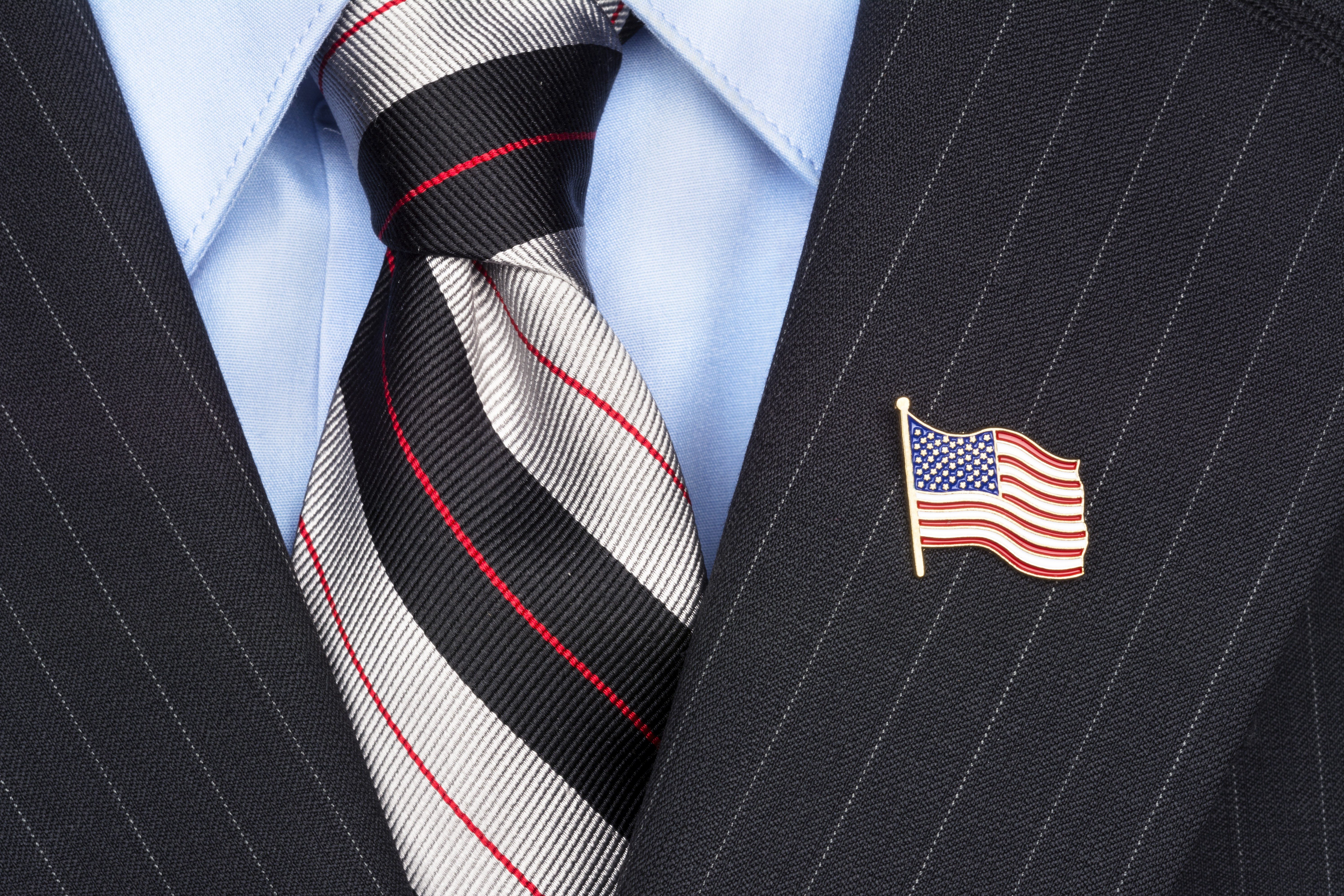 A striped necktie rests on a blue shirt and pinstriped suit, adorned with an American flag pin.