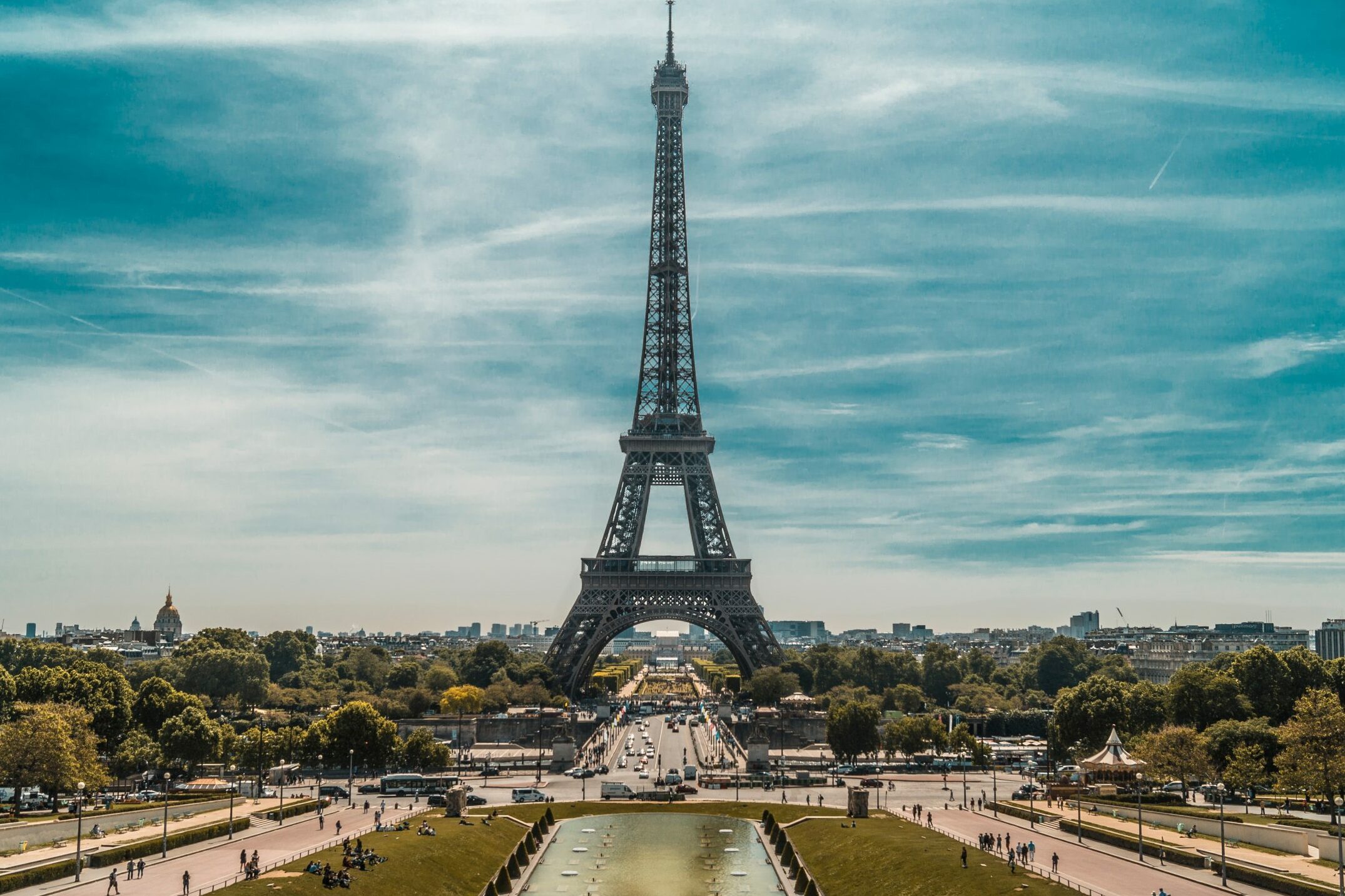 The Eiffel Tower stands tall, dominating the skyline amid scattered clouds, with people walking and relaxing in a nearby park.