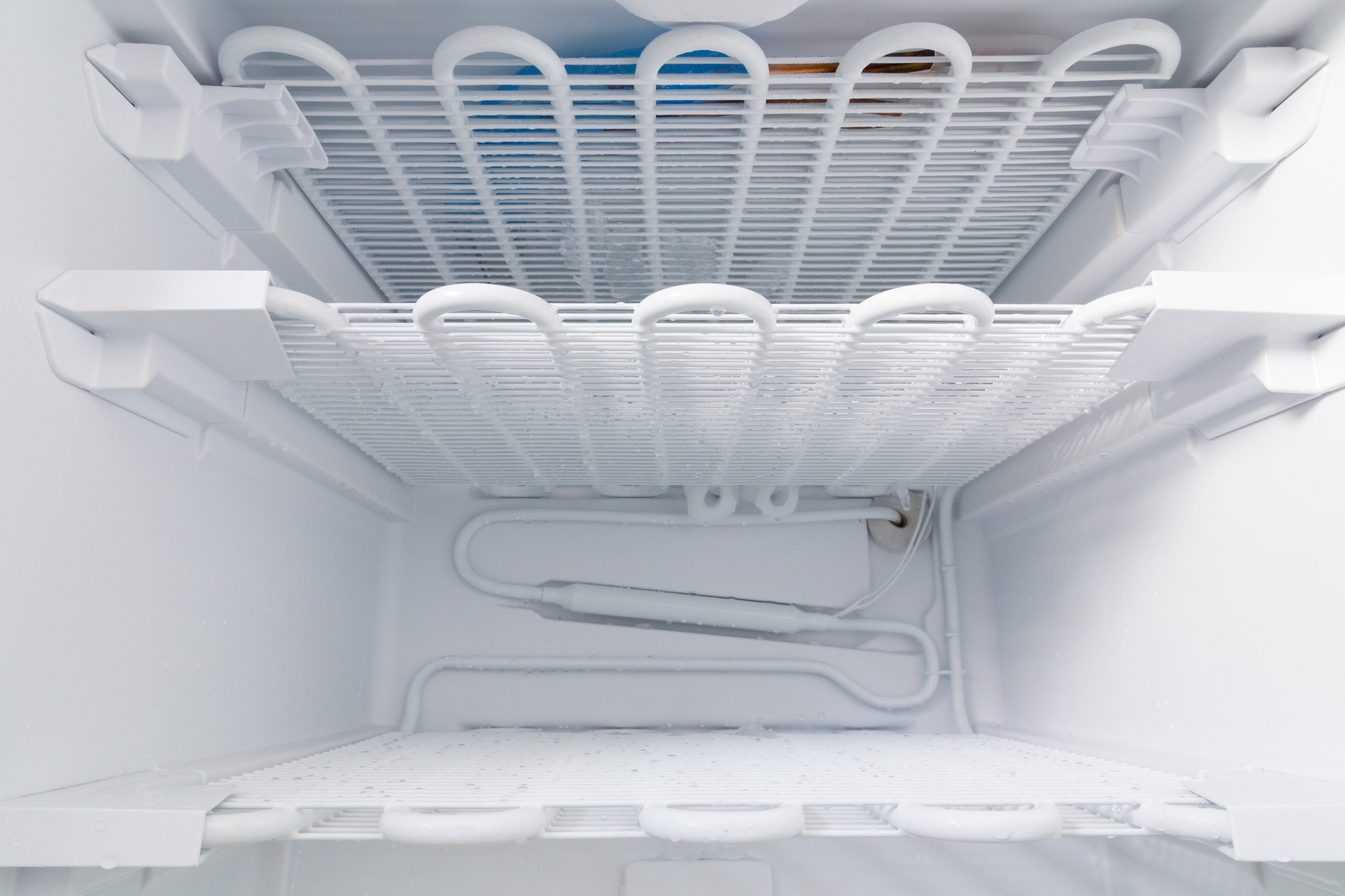 Empty freezer shelves covered with frost and ice, enclosed within a refrigerator interior, showcasing plumbing and cooling elements.