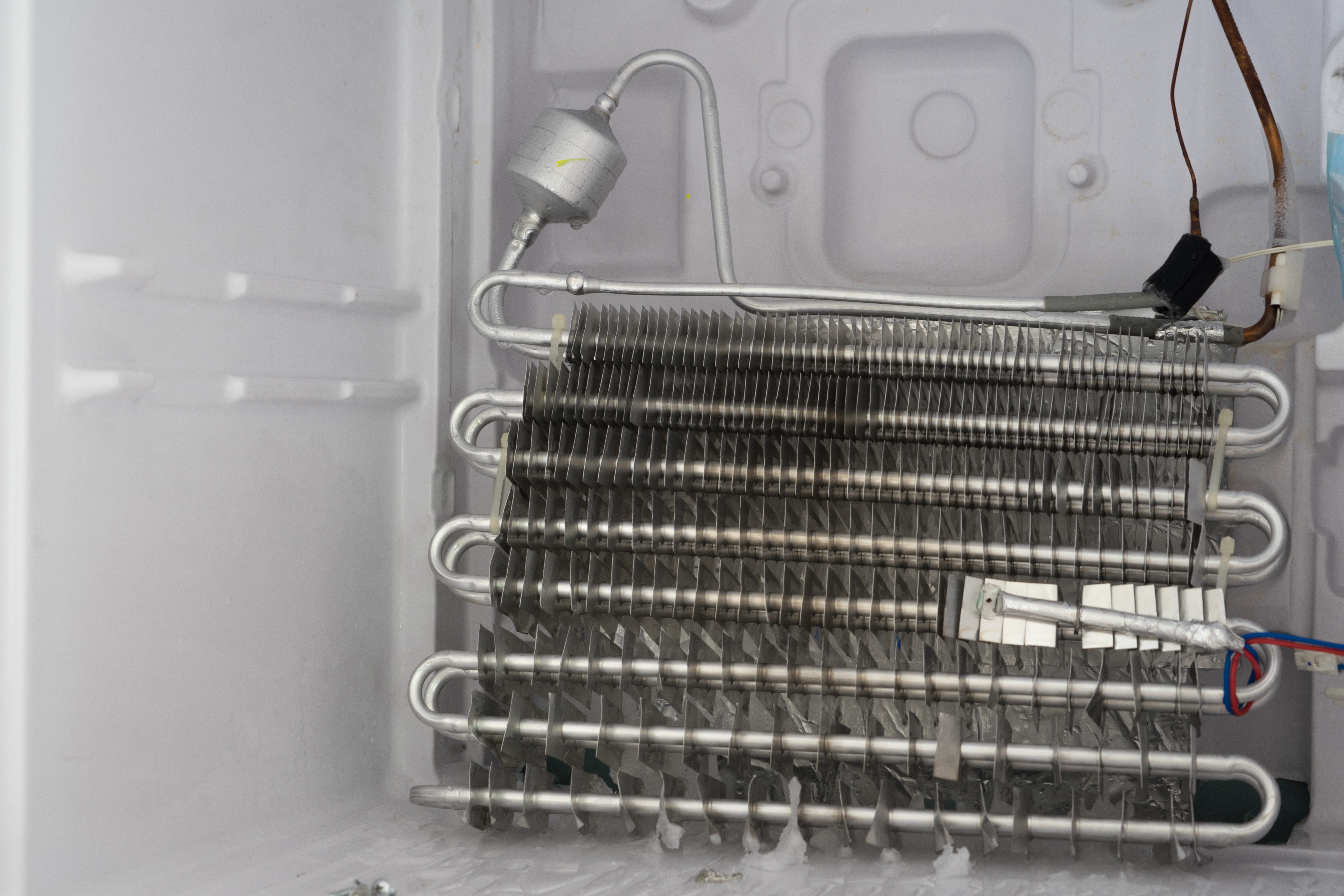 Metallic coils lay horizontally, connected and attached inside an open freezer compartment with visible frost on the coils and walls.