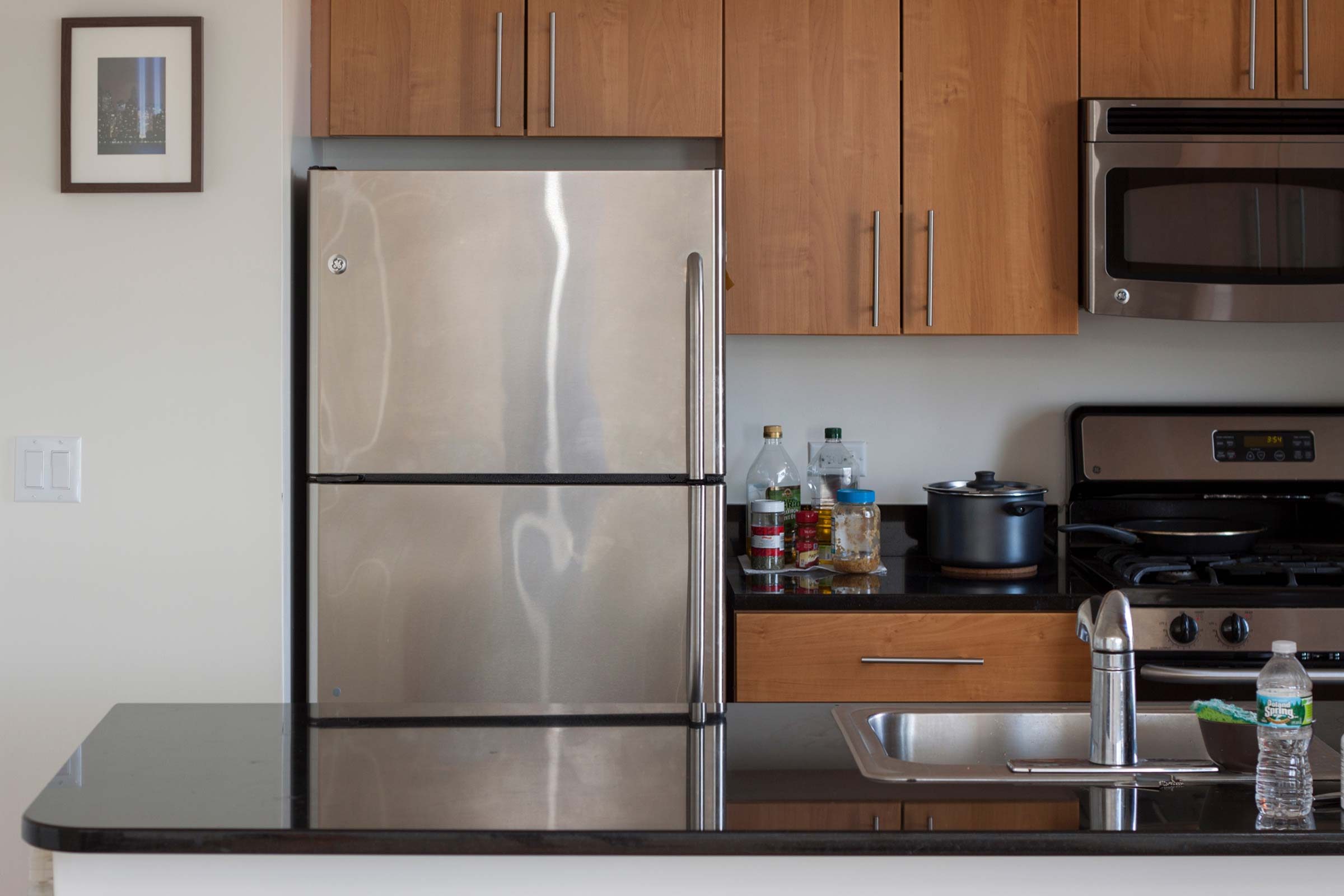 Stainless steel refrigerator stands between wooden cabinets in a kitchen with a stove, countertop, sink, and miscellaneous bottles nearby.
