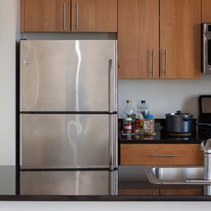 Stainless steel refrigerator stands between wooden cabinets in a kitchen with a stove, countertop, sink, and miscellaneous bottles nearby.
