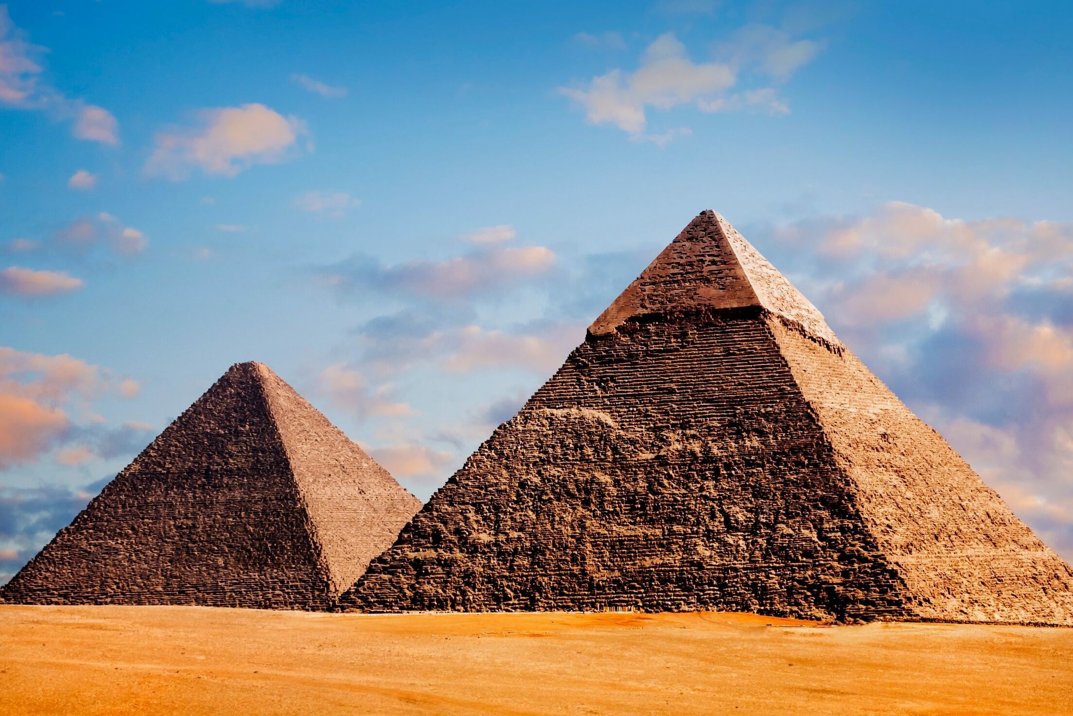 Pyramids stand solitary under a blue sky with scattered clouds. Sandy desert surrounds the ancient structures, evoking a timeless atmosphere.
