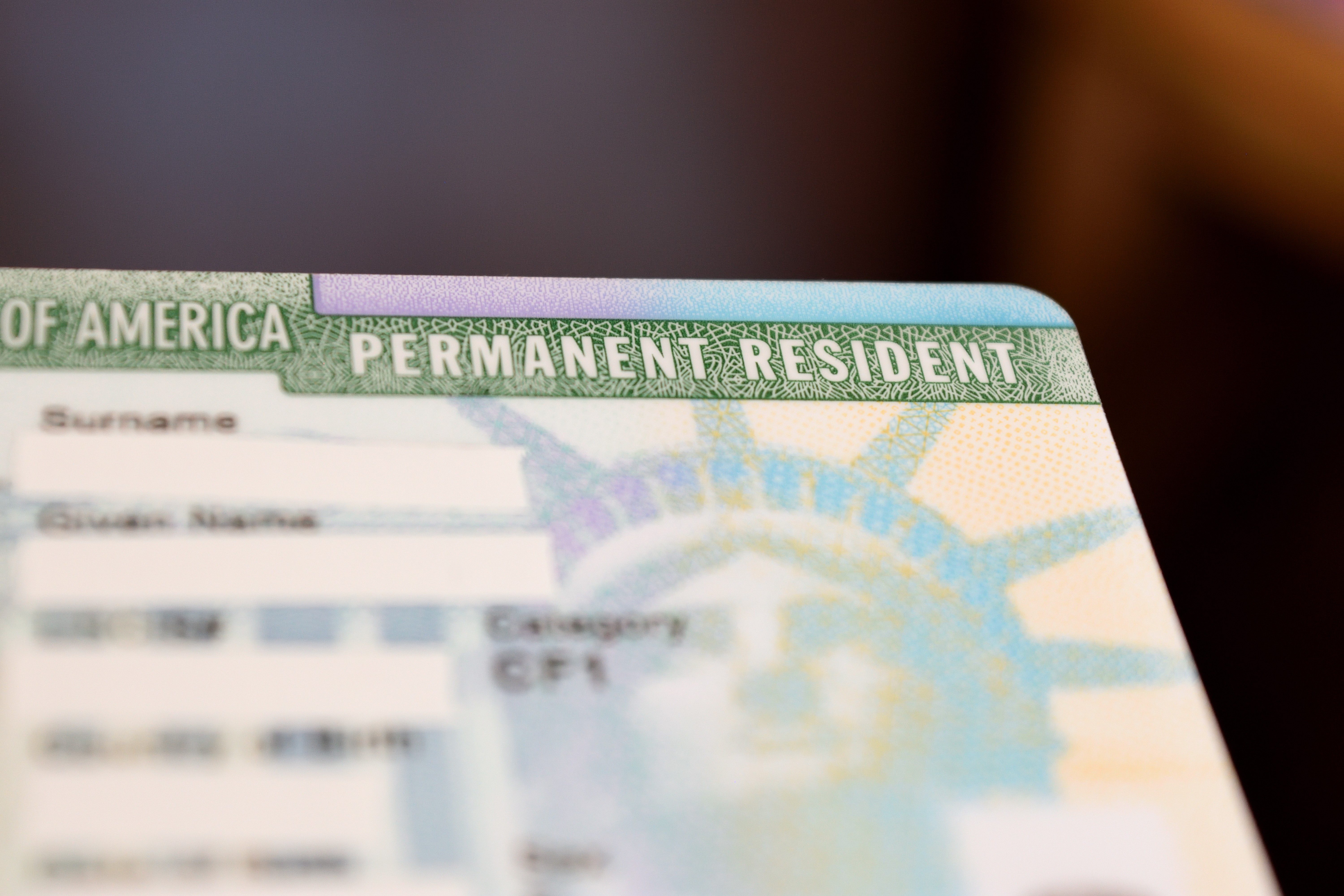 A U.S. green card with partially visible text "PERMANENT RESIDENT," featuring a blurry background and limited personal information areas.