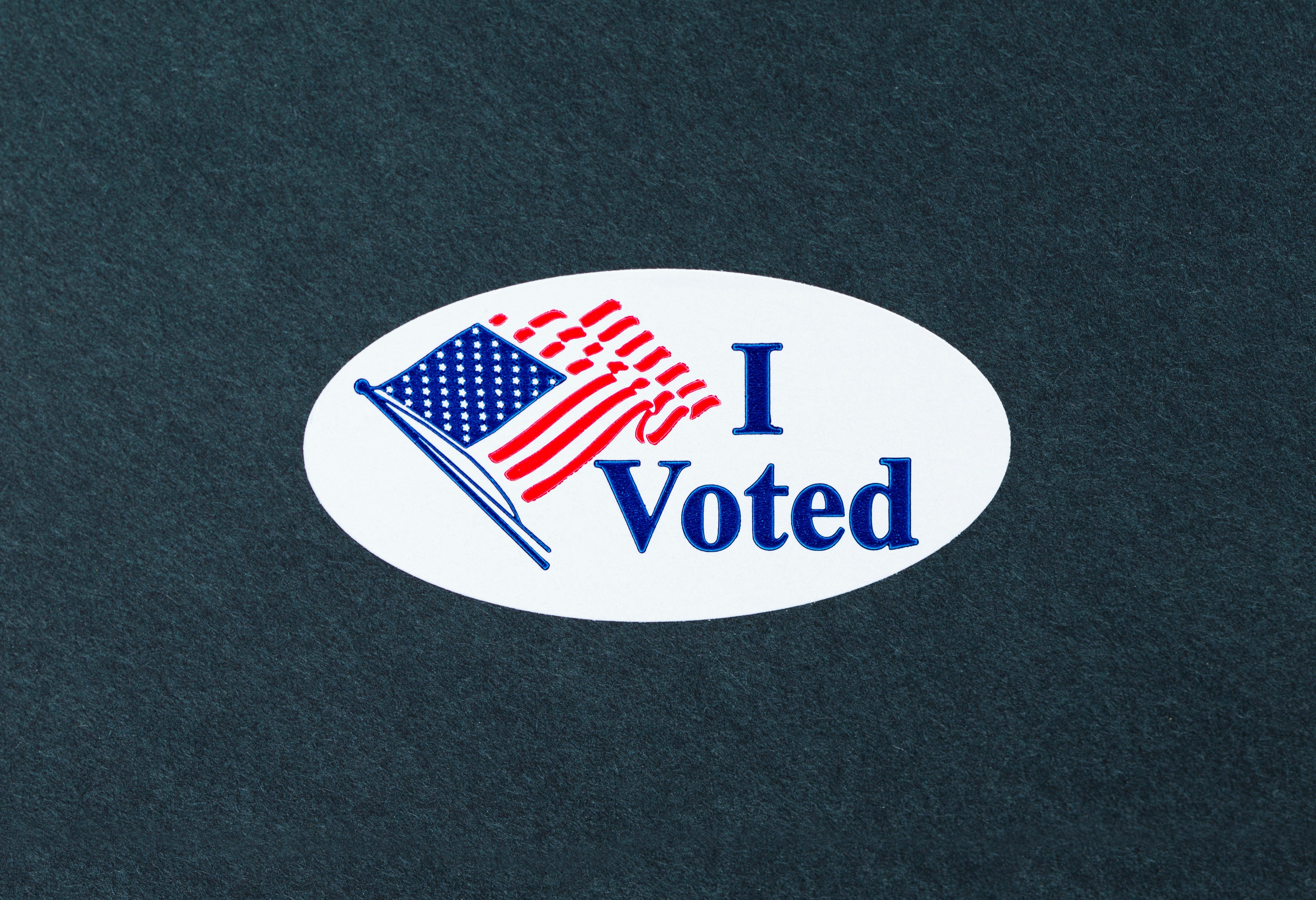 Sticker displays the text "I Voted" with an American flag graphic, placed on a dark textured background.