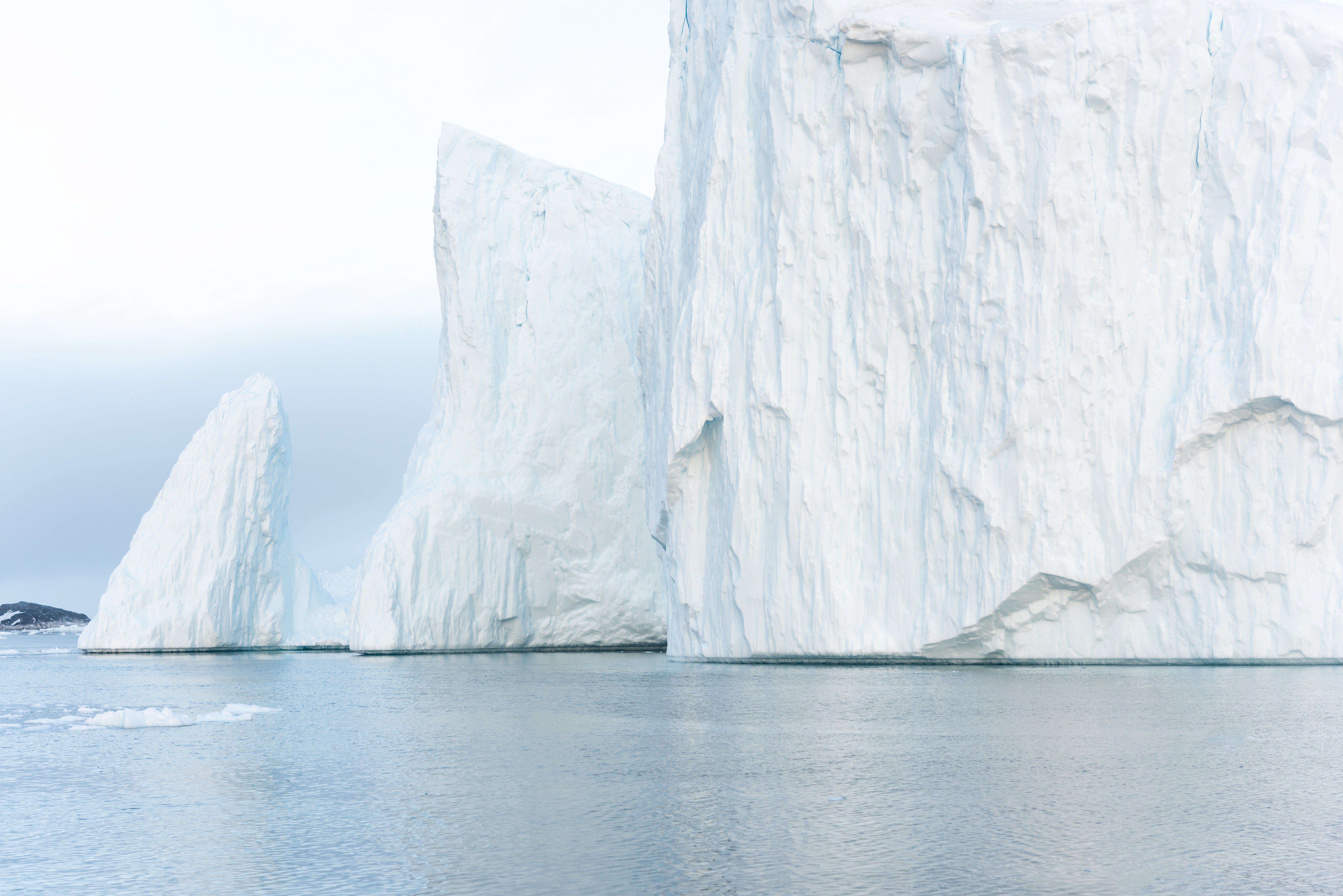 Icebergs tower above still ocean waters; icy, calm seascape under overcast skies.