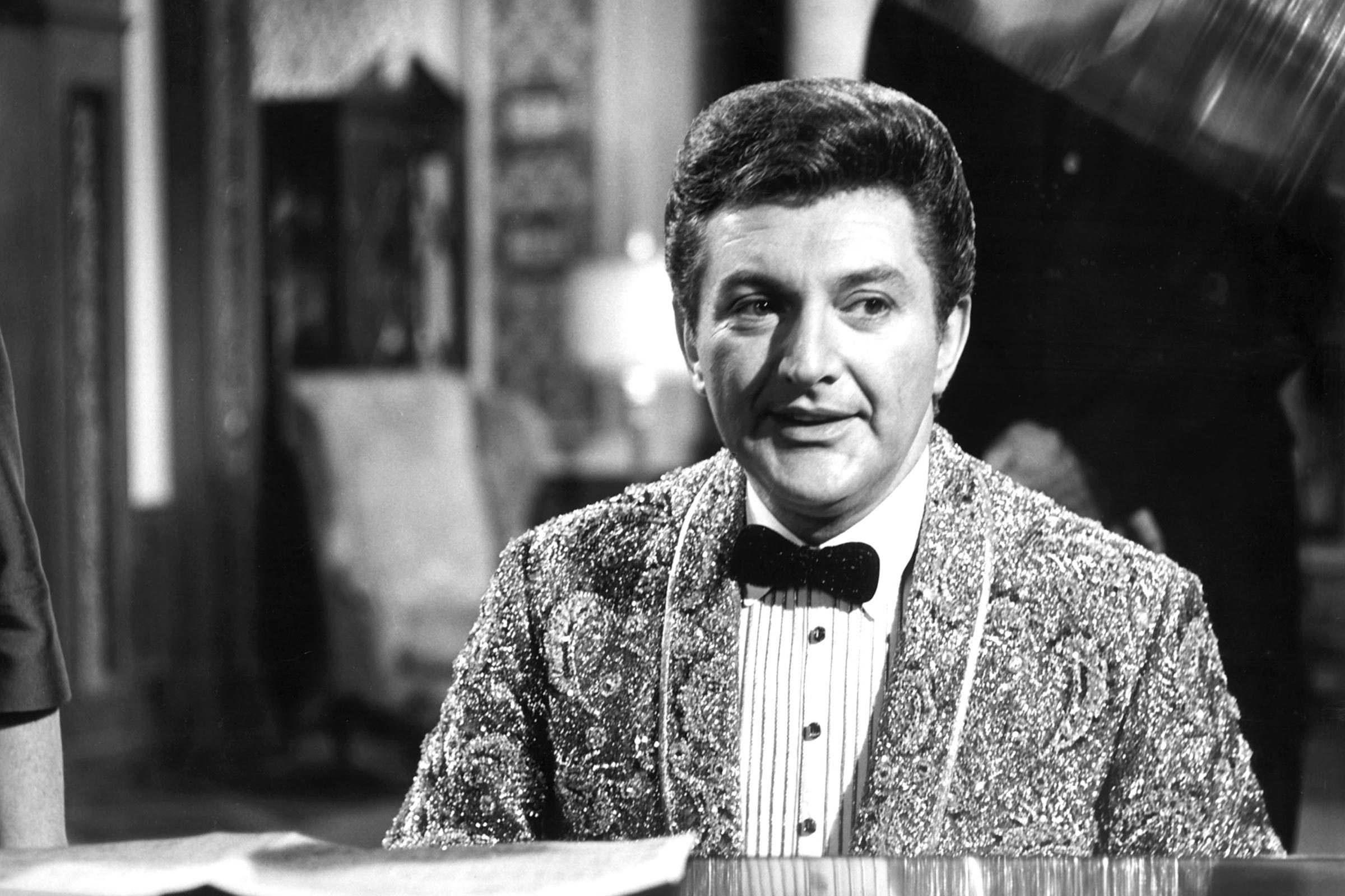 Man sits at a piano, wearing a sequined jacket, in an ornate room with furniture and decorative wallpaper.