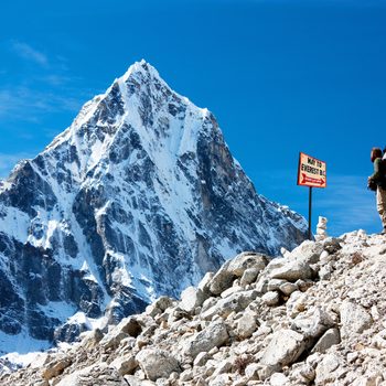 Hiker stands observing snowy mountain peak; sign reads "Way to Everest B.C." in rocky, mountainous terrain under clear blue sky.