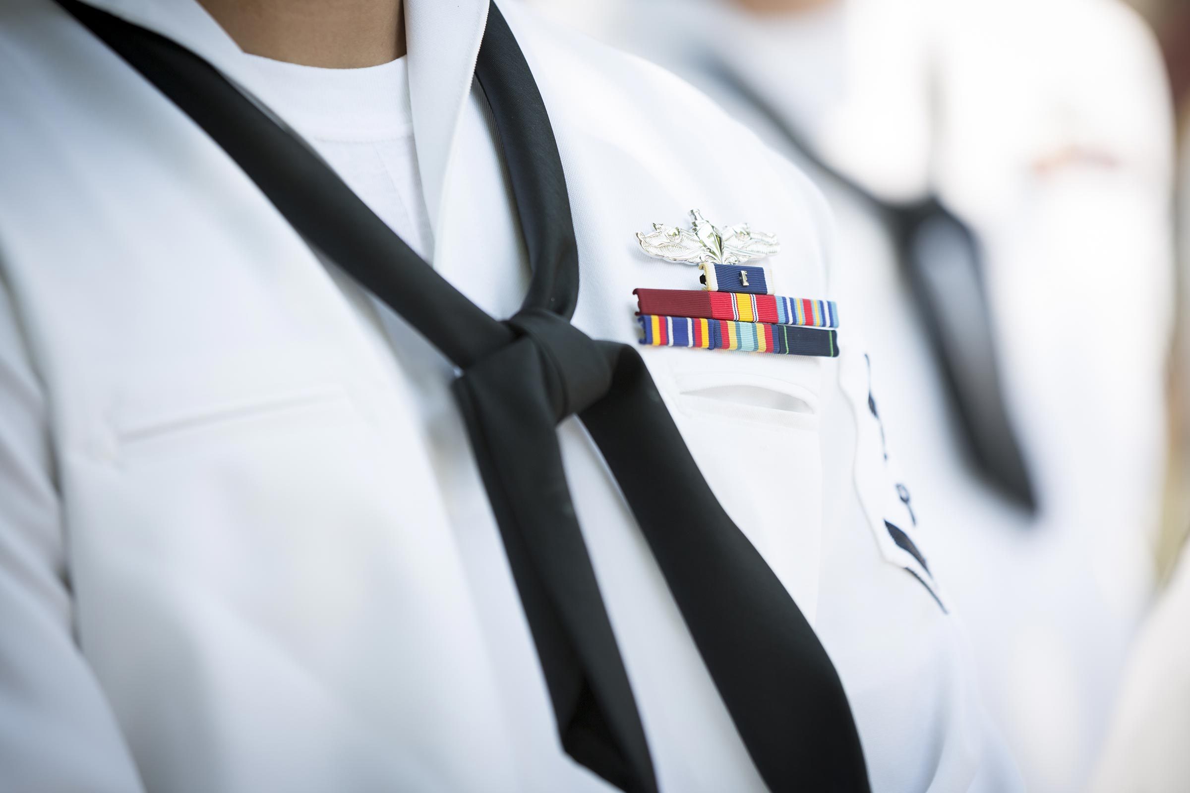 A person wears a white naval uniform adorned with colorful ribbons and a black necktie, standing in a formal setting.