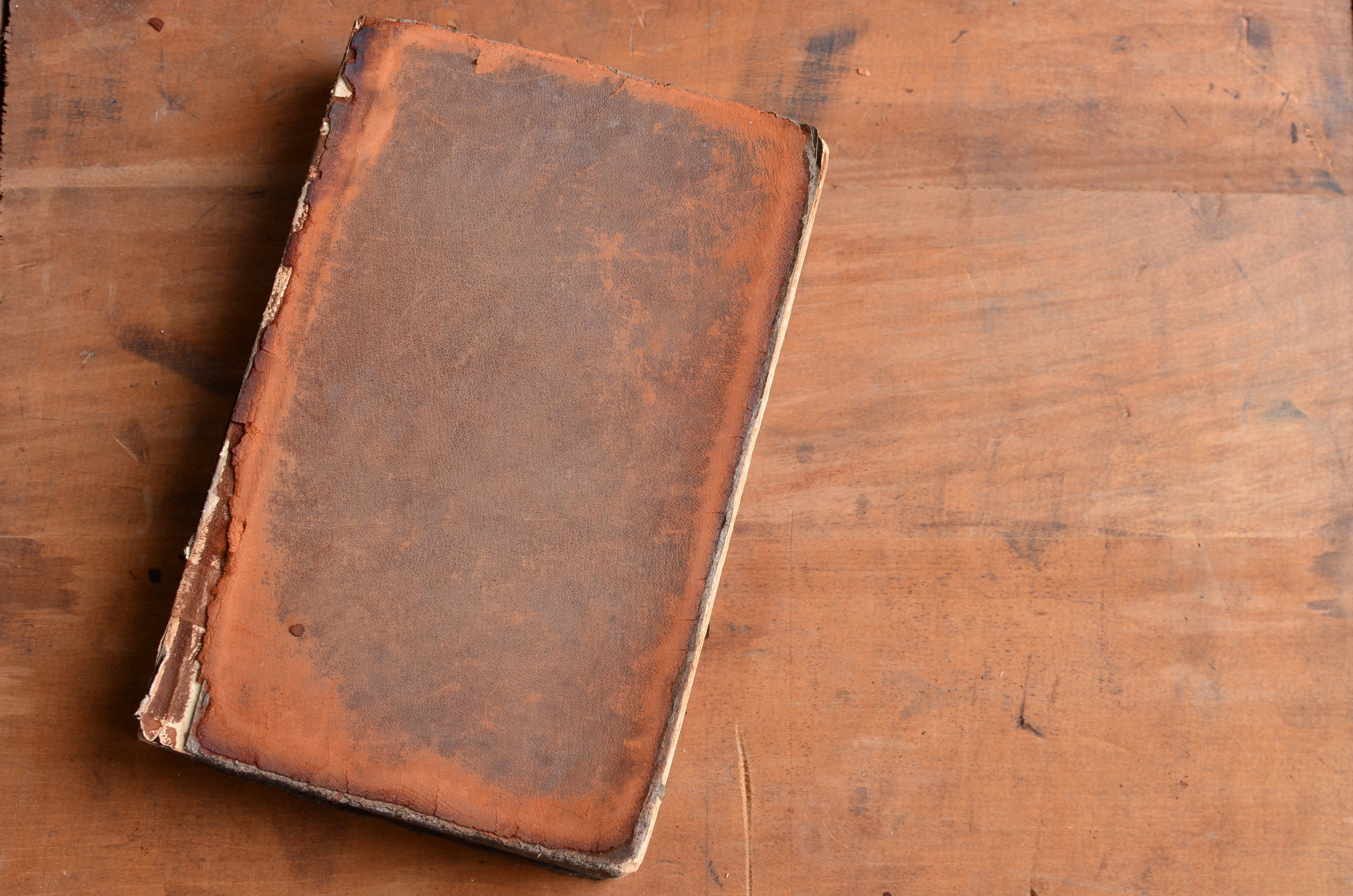 A worn, old book rests closed on a wooden table, highlighting its aged, textured leather cover.