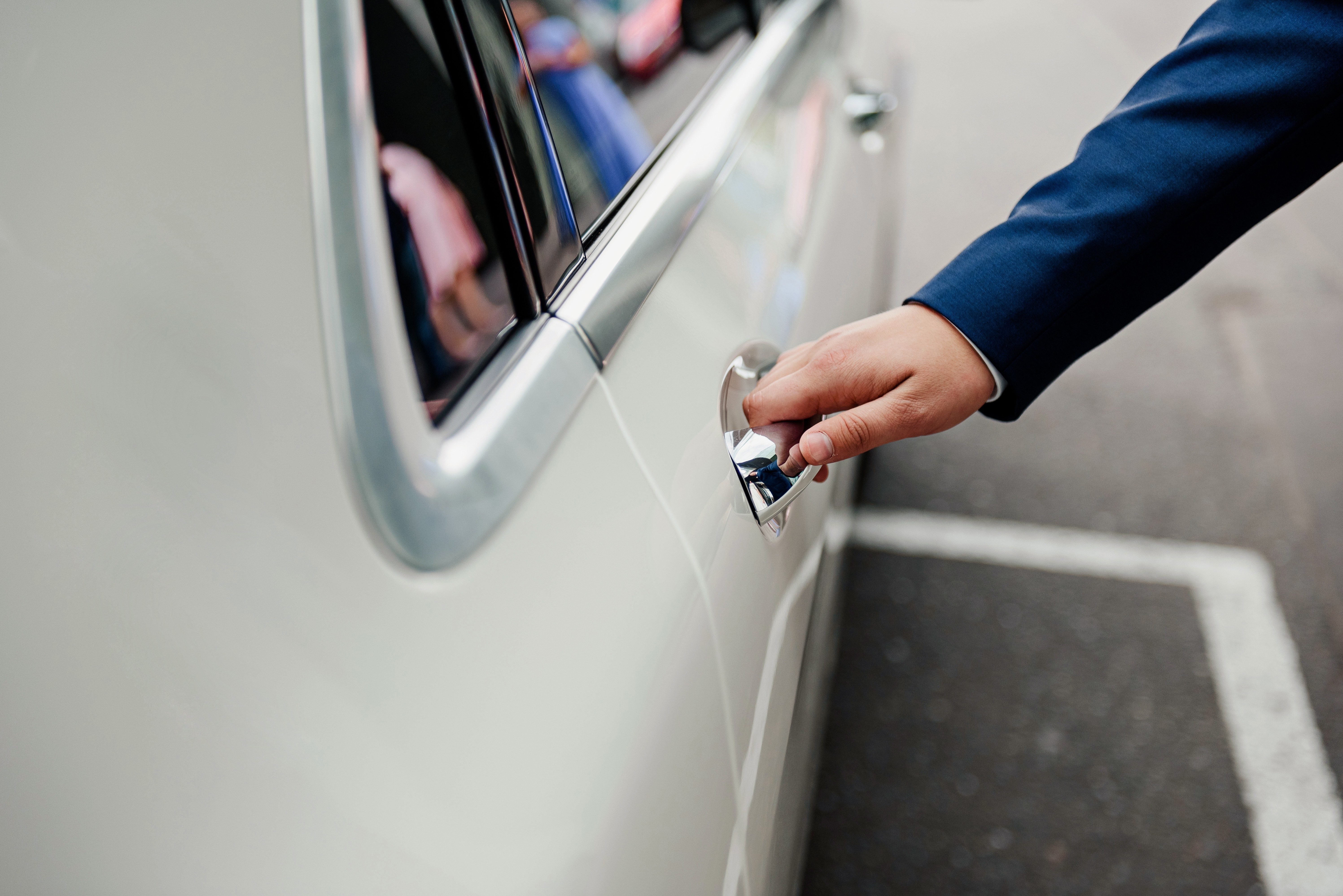 Hand opening a car door, wearing a blue suit, in a parking lot setting.