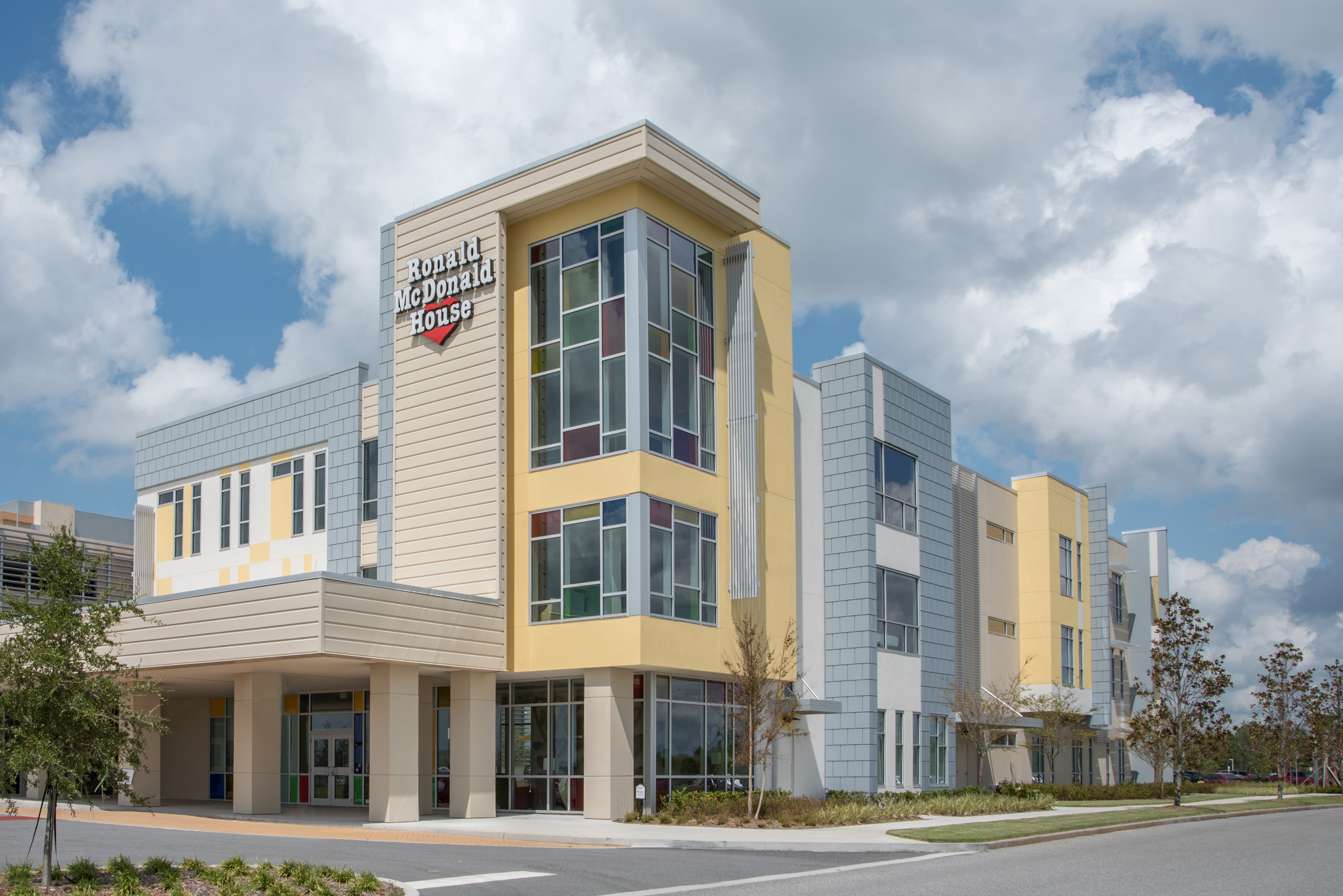 Modern building with colorful windows stands under a partly cloudy sky, surrounded by trees and a street. Text reads: "Ronald McDonald House."