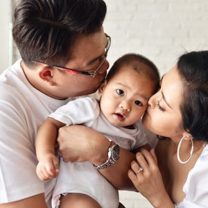 Baby receives kisses from two adults, in a white brick room.
