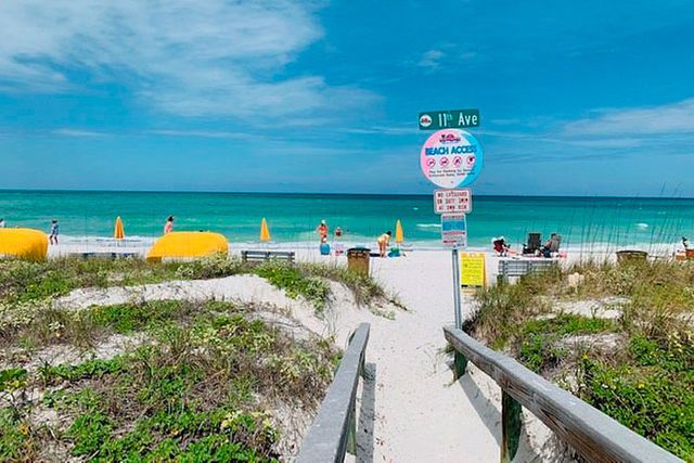 Pathway leads to beach; people sunbathe near yellow umbrellas. Signs indicate beach access. Lush greenery surrounds sandy path; ocean waves and blue sky beyond.