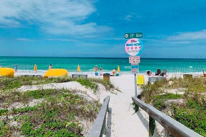 Pathway leads to beach; people sunbathe near yellow umbrellas. Signs indicate beach access. Lush greenery surrounds sandy path; ocean waves and blue sky beyond.