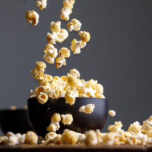 Popcorn falls into a black bowl, surrounded by scattered popcorn on a wooden surface, set against a dark background.