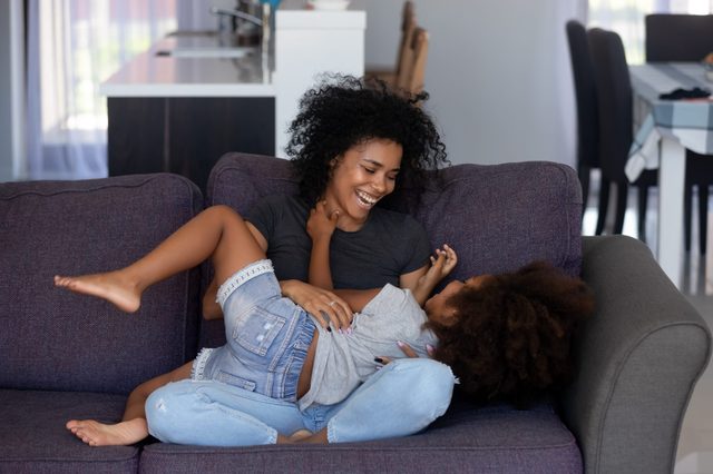 Smiling woman playfully tickles a child on a purple couch in a cozy living room with a dining area in the background.