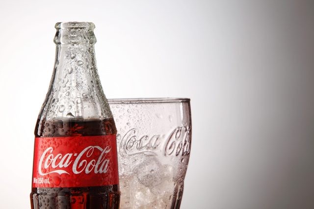 A Coca-Cola bottle, covered in condensation, stands beside a glass with ice on a plain background.