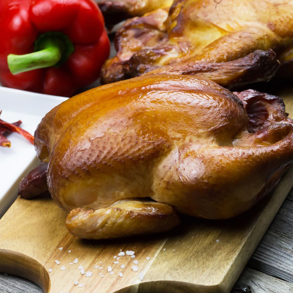 Roasted chicken sits on a wooden board with salt, accompanied by a red bell pepper and dried chili peppers in the background.