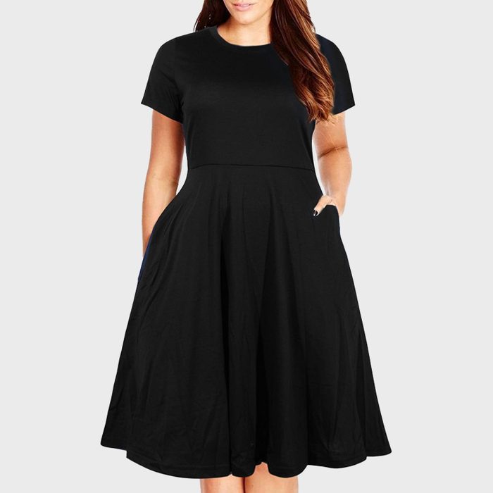 A person stands wearing a knee-length black dress with short sleeves, hands in pockets, on a plain gray background.