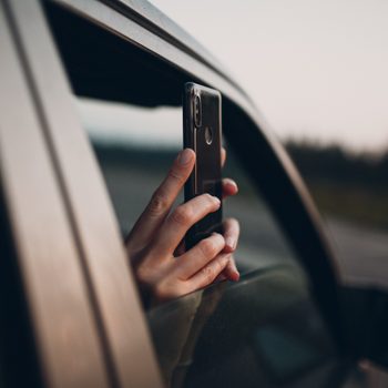 Hand holds a smartphone out of a car window, capturing a photograph, with a blurred outdoor landscape in the background.
