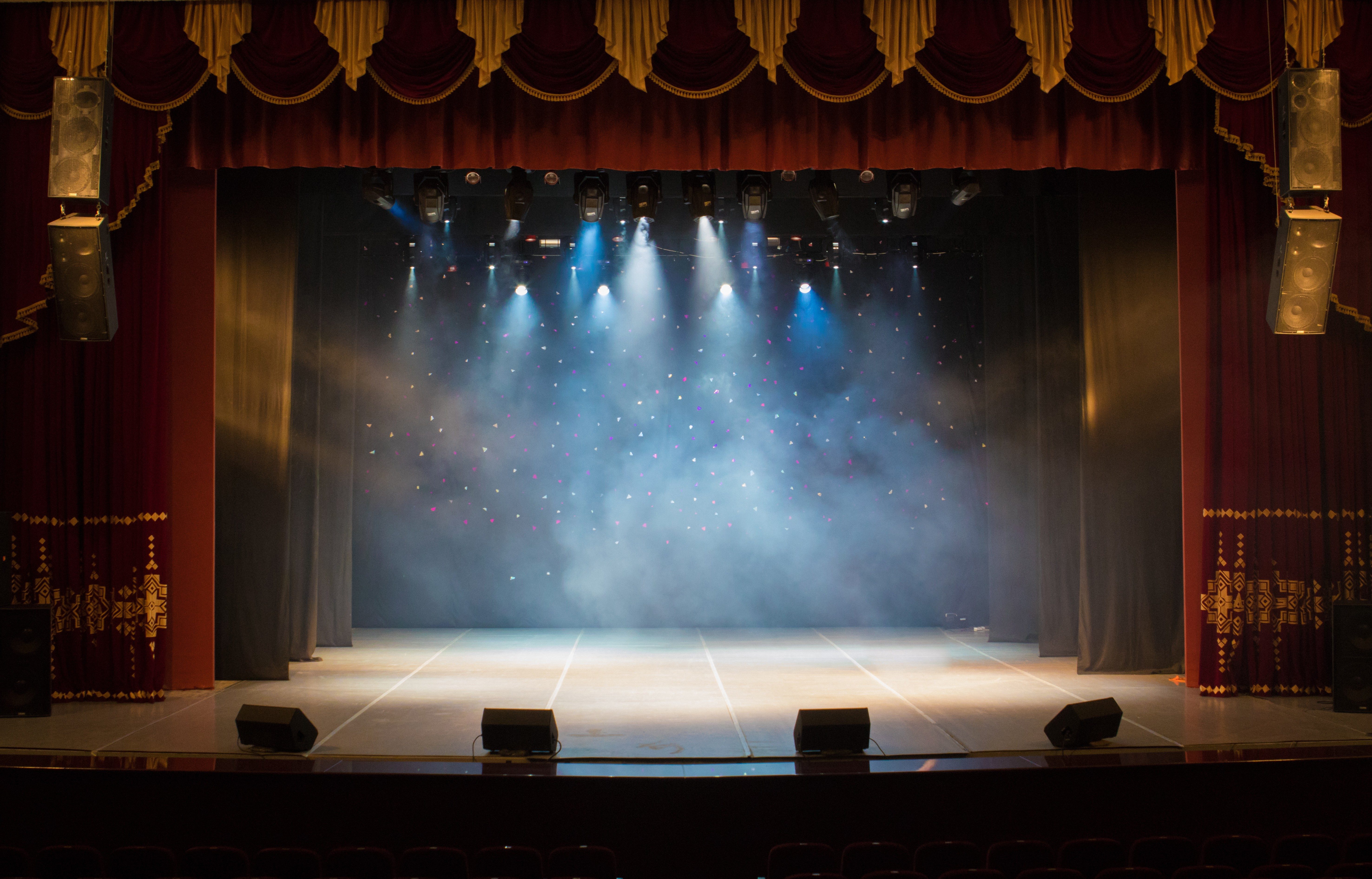 Stage emits fog under blue spotlights, surrounded by dark red curtains and background of tiny colorful lights.