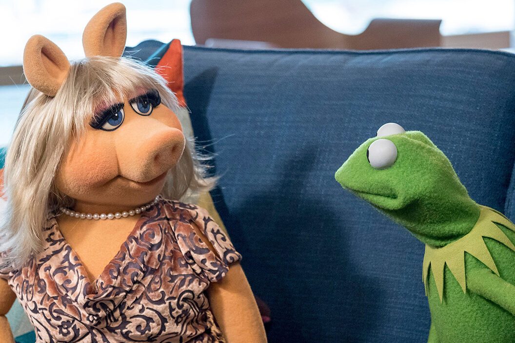 Two puppet characters sit conversing on a blue couch, in a brightly lit room with patterned cushions.