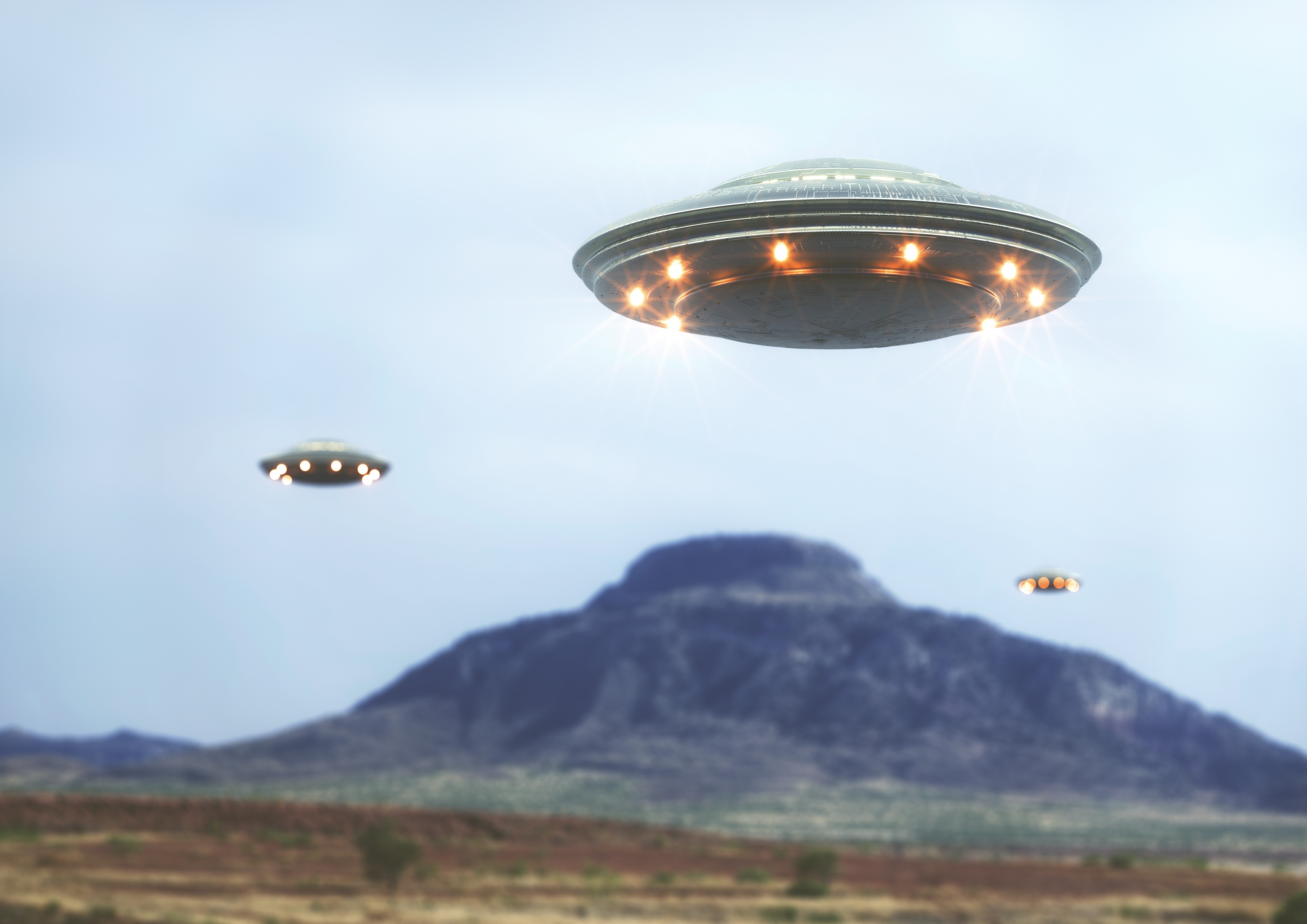 Three flying saucers hover above a mountainous landscape, glowing lights beneath them, in a cloudy sky.