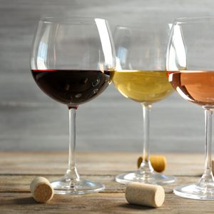 Three wine glasses filled with red, white, and rosÃ© wine sit on a wooden table with corks scattered around against a neutral background.