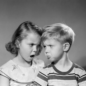 A young girl and boy, both with pouting expressions, stand against a plain background, appearing to disagree or argue.
