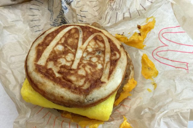 Breakfast sandwich displaying browned top bun with "M" logo, rests on crumpled wrapper. Melted cheese and folded egg peeking out underneath.