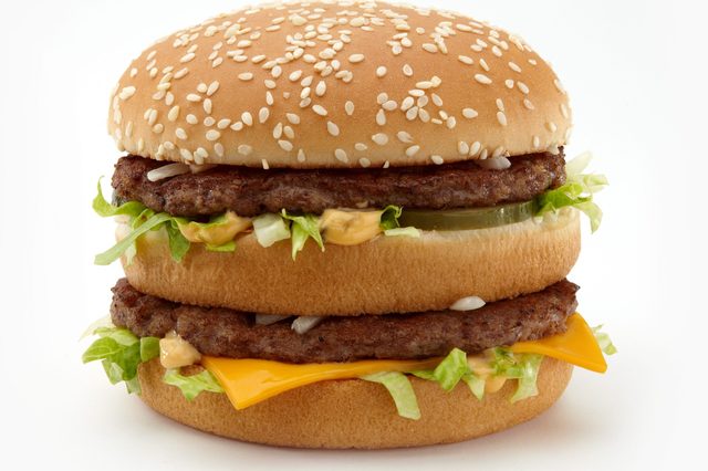 A sesame seed burger with two beef patties and cheese rests between layers of lettuce, pickles, and sauce on a plain background.