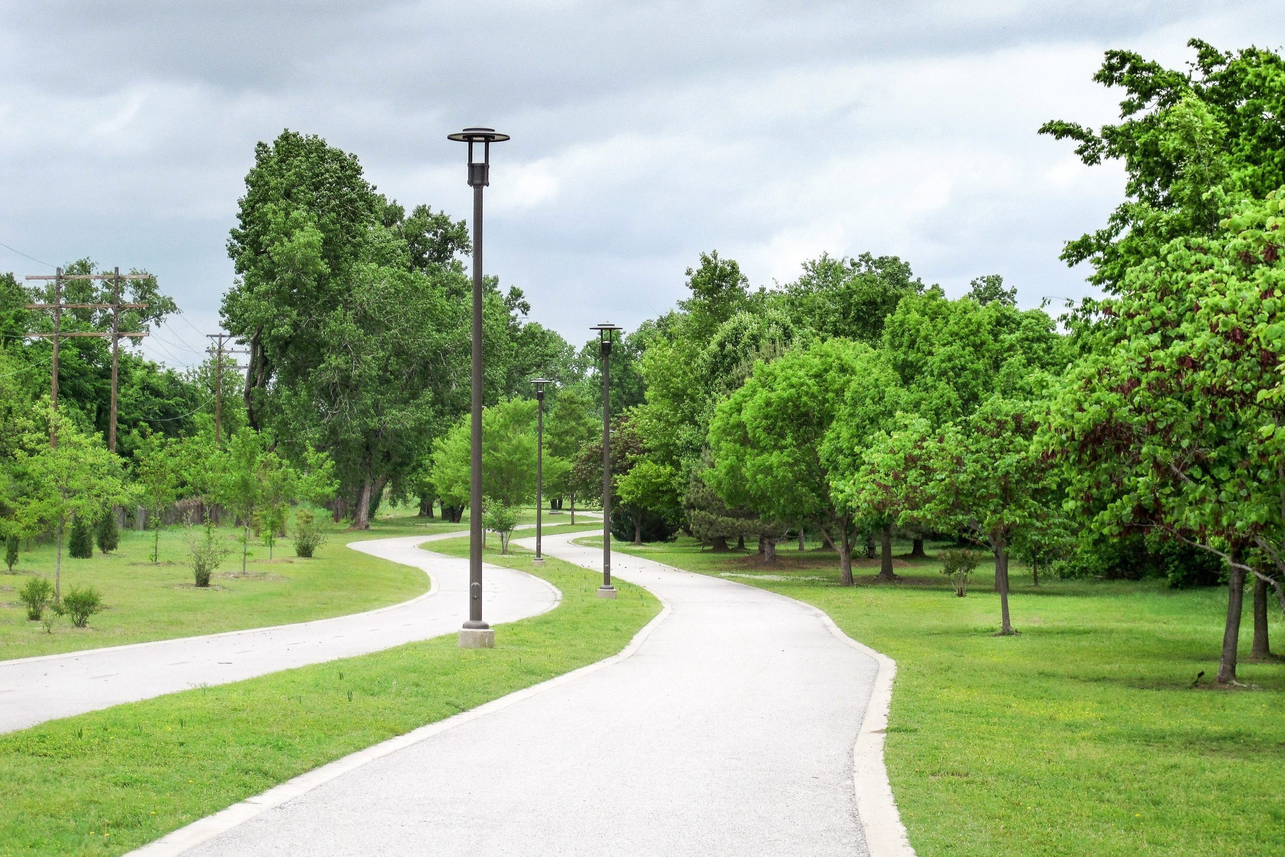 Path curves through a park, flanked by green trees and lamp posts, under a cloudy sky.