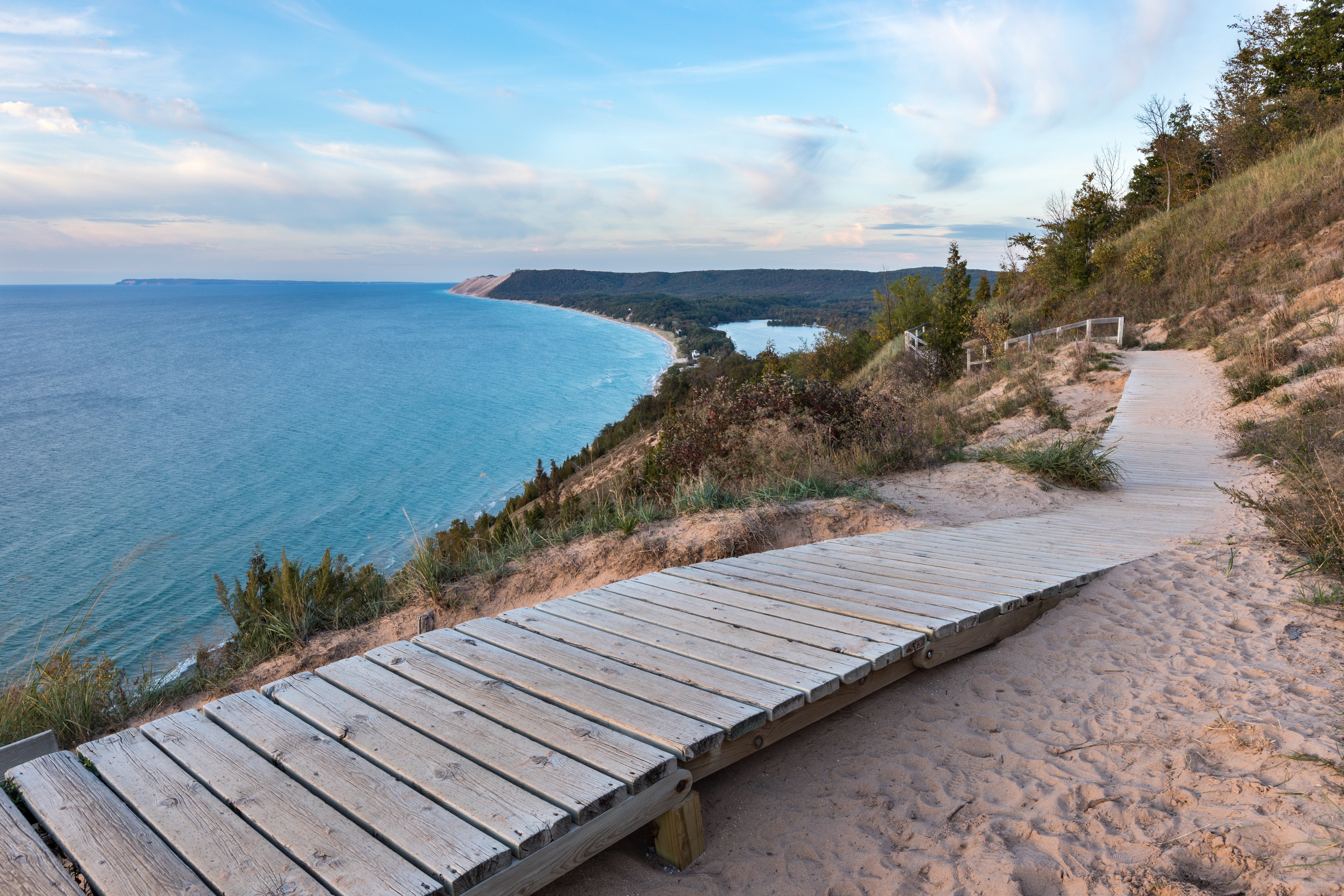 A wooden boardwalk winds gently through sandy dunes overlooking a vast, calm blue ocean under a clear sky.