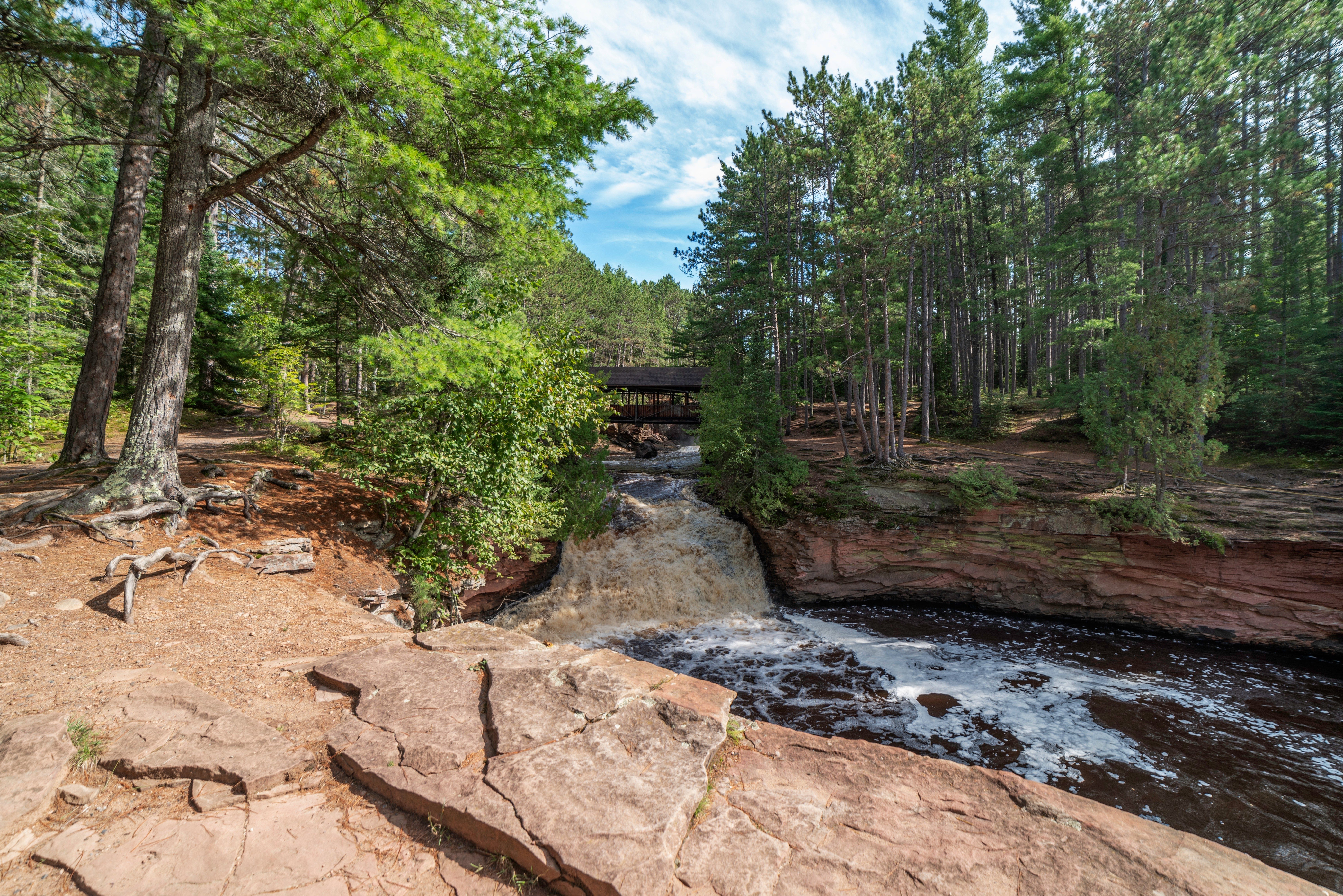 Waterfall cascades over rocky ledge, surrounded by dense evergreen forest under a bright blue sky.