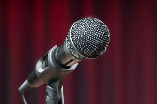 Microphone angled towards an audience, set against a blurred red curtain backdrop.