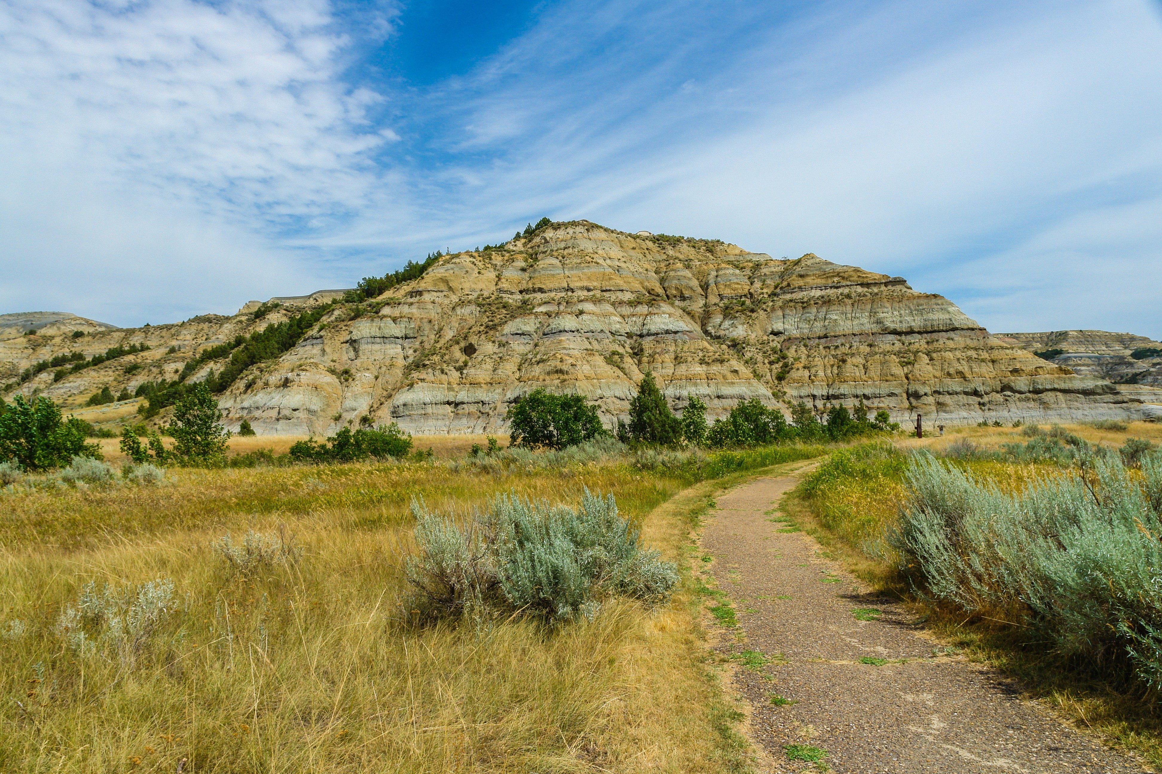 A winding dirt path leads through grassy fields toward layered rocky hills under a partly cloudy sky.