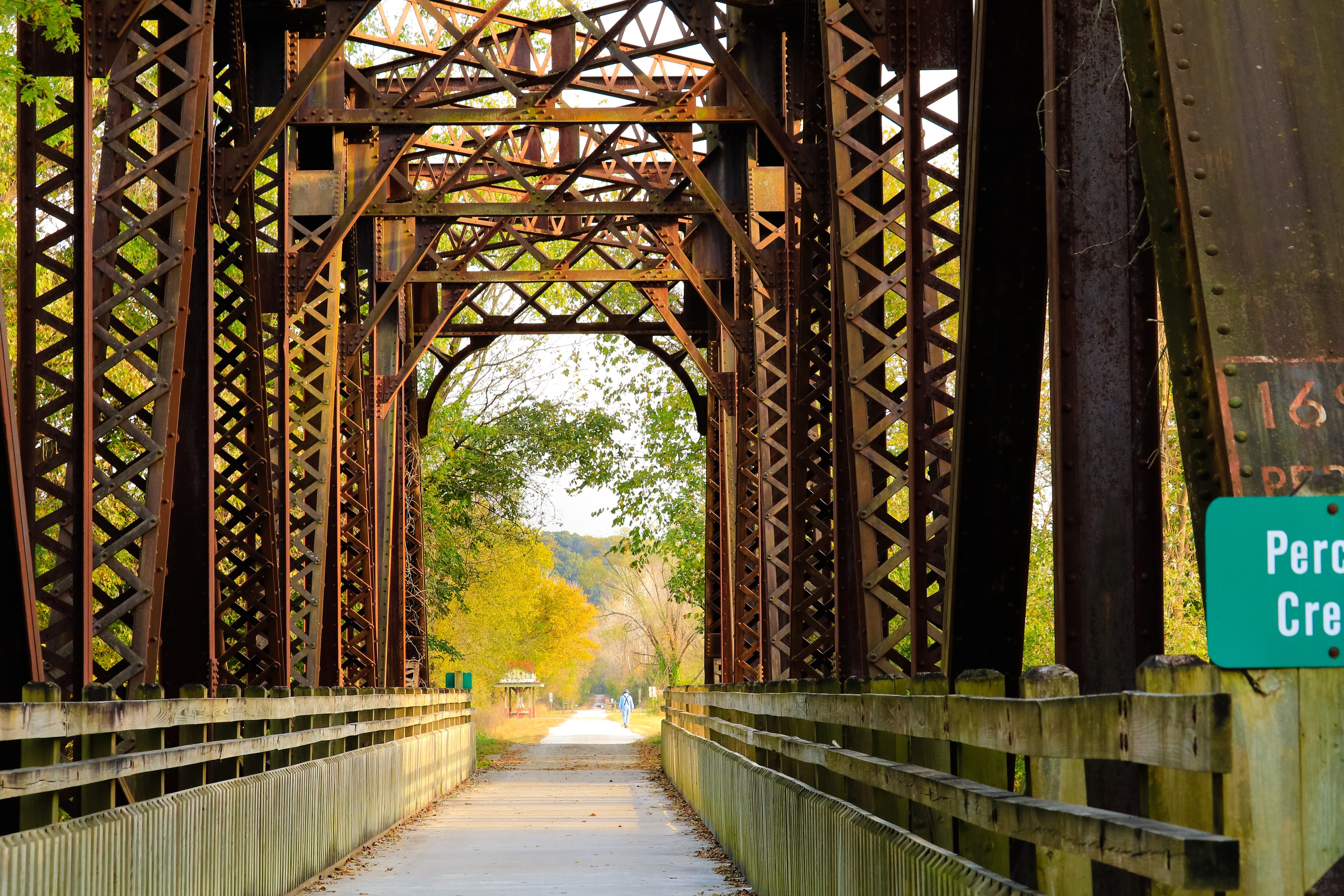 Rusty metal bridge stretches over a path, surrounded by trees, with a sign reading "Perch Creek Trail" to the side.