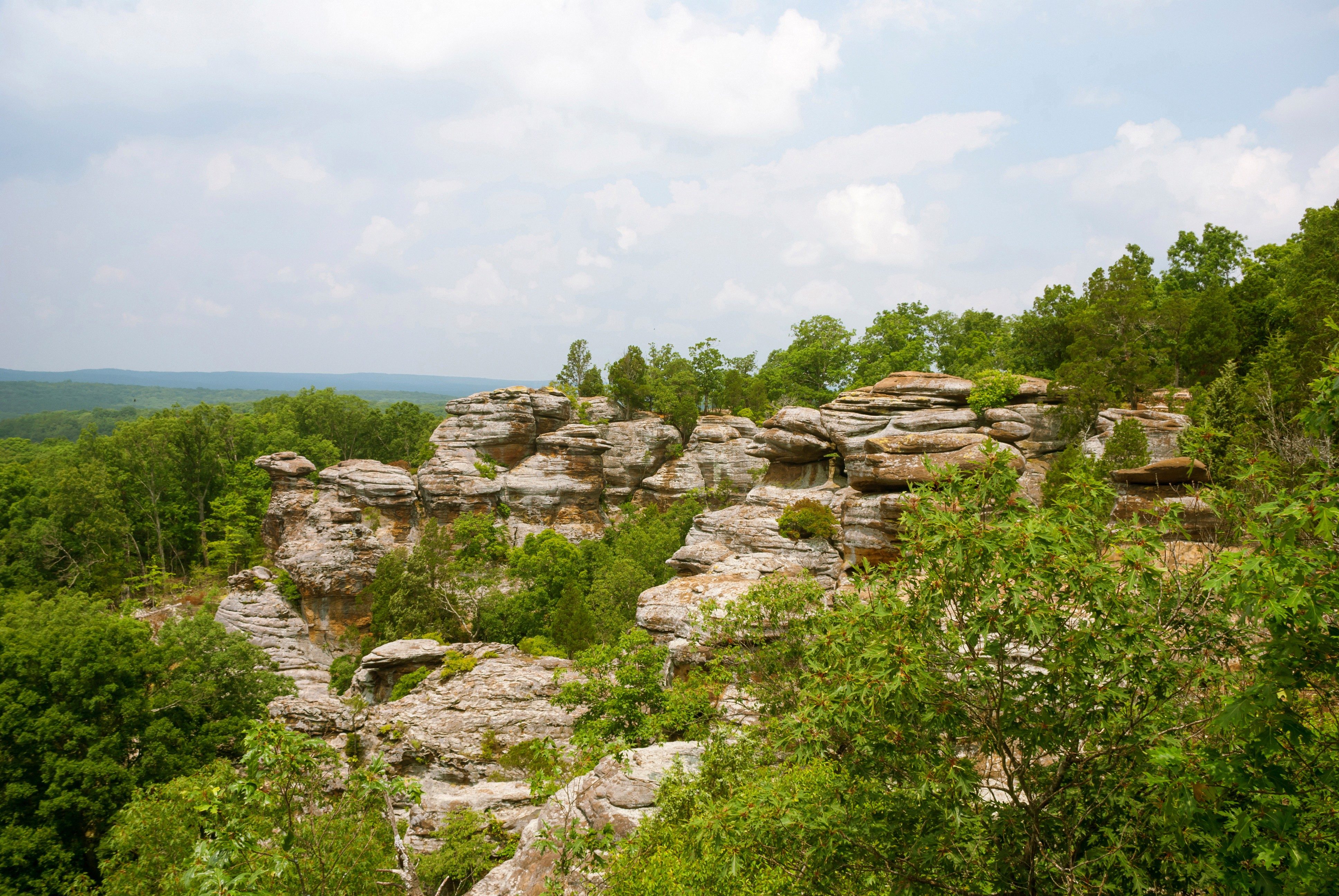 Rock formations stand prominently amidst lush green trees under a partly cloudy sky, creating a picturesque natural landscape.