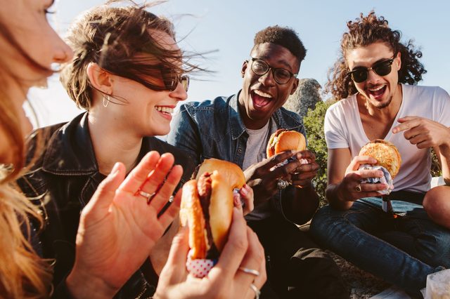 People laughing and eating burgers outdoors, enjoying a sunny day with trees and rocks in the background.