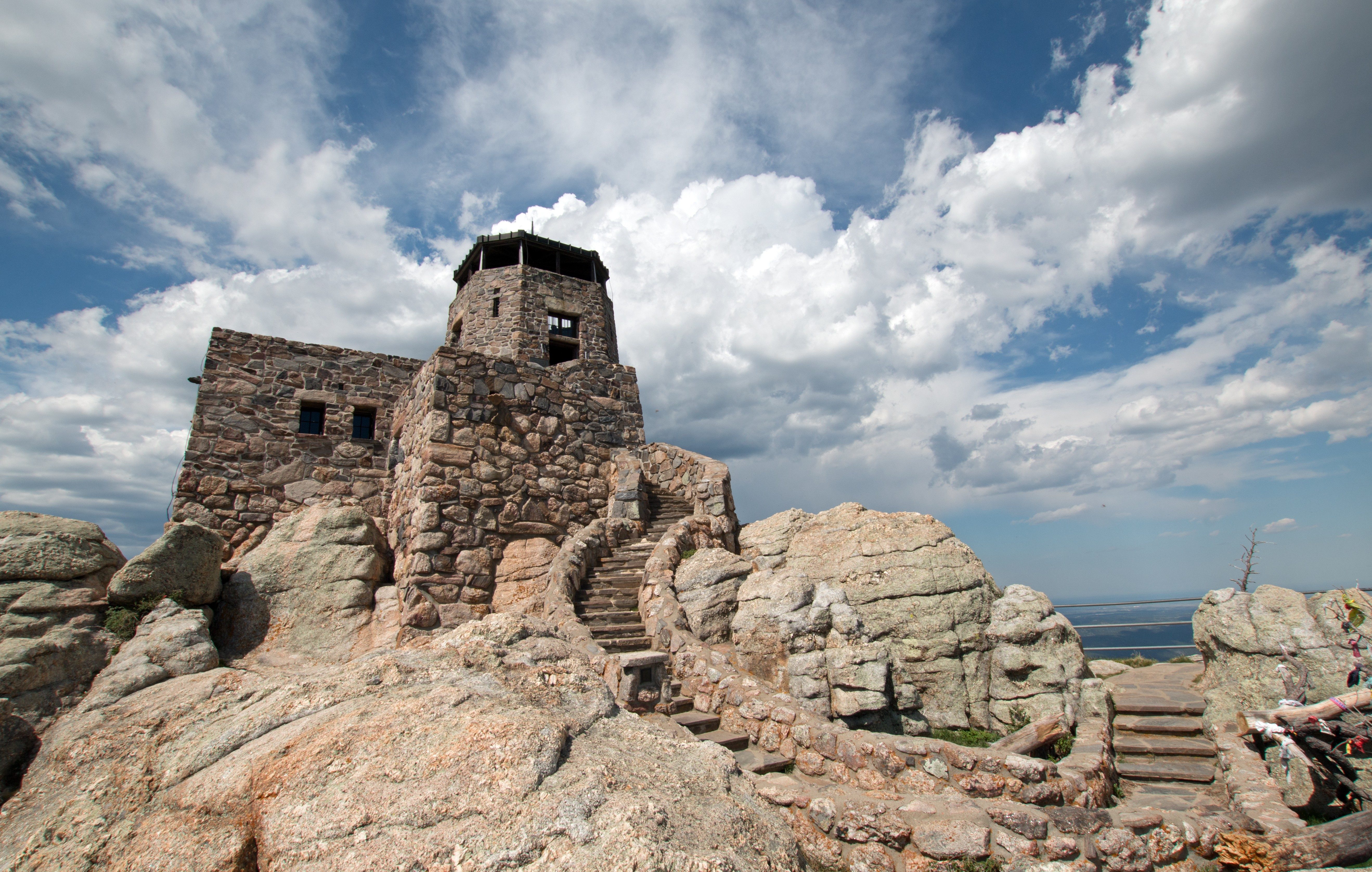 Stone lookout tower sits atop rocky terrain under a partly cloudy sky, with curved stone steps ascending to it.
