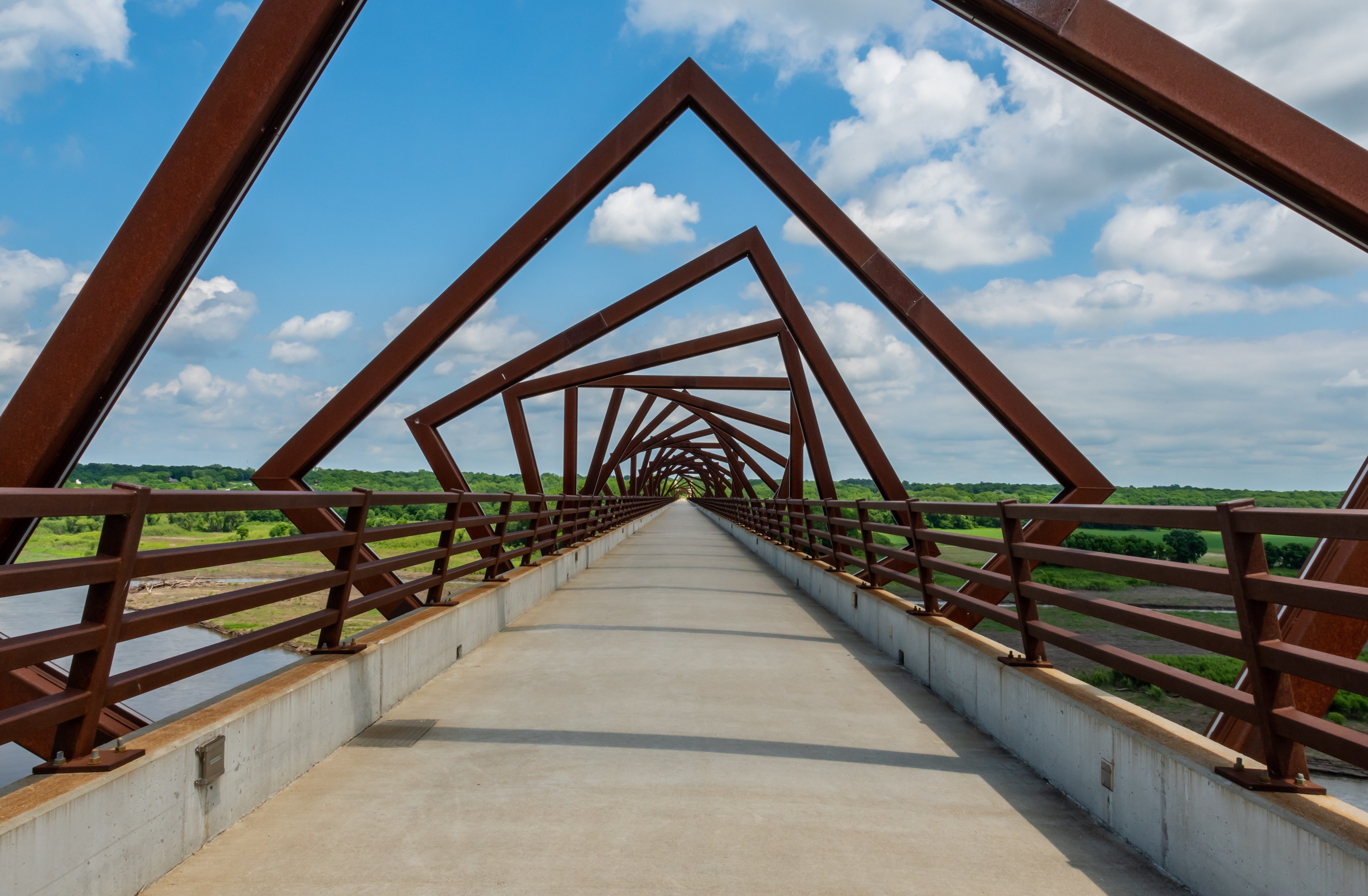 Geometric bridge structure forms repeating triangular patterns, spanning across a wide river with lush green landscapes under a clear blue sky.