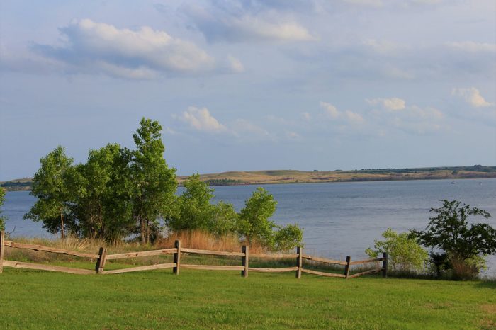 A shot of Kanopolis Lake on the north side of a Wooden Fence, green grass,water,trees, blue sky and clouds, thats bright and colorful.