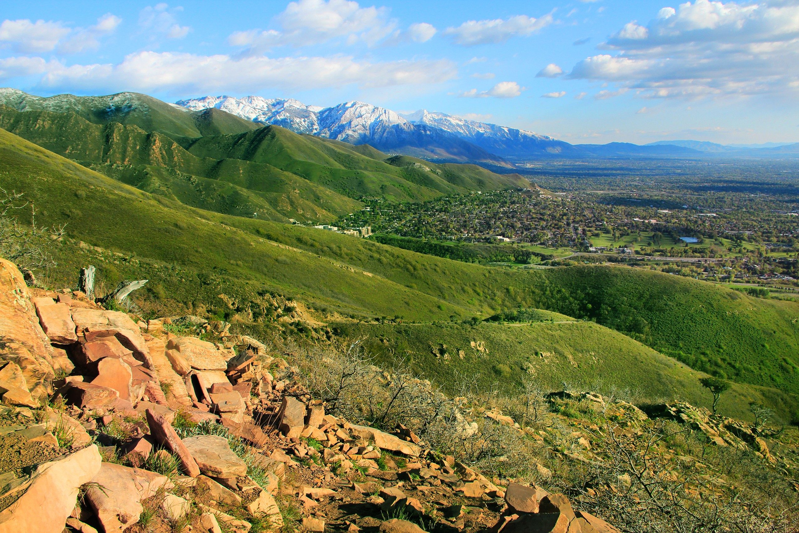 Rocky foreground leads to green hills extending towards distant snow-capped mountains under a partly cloudy sky with a town nestled below.